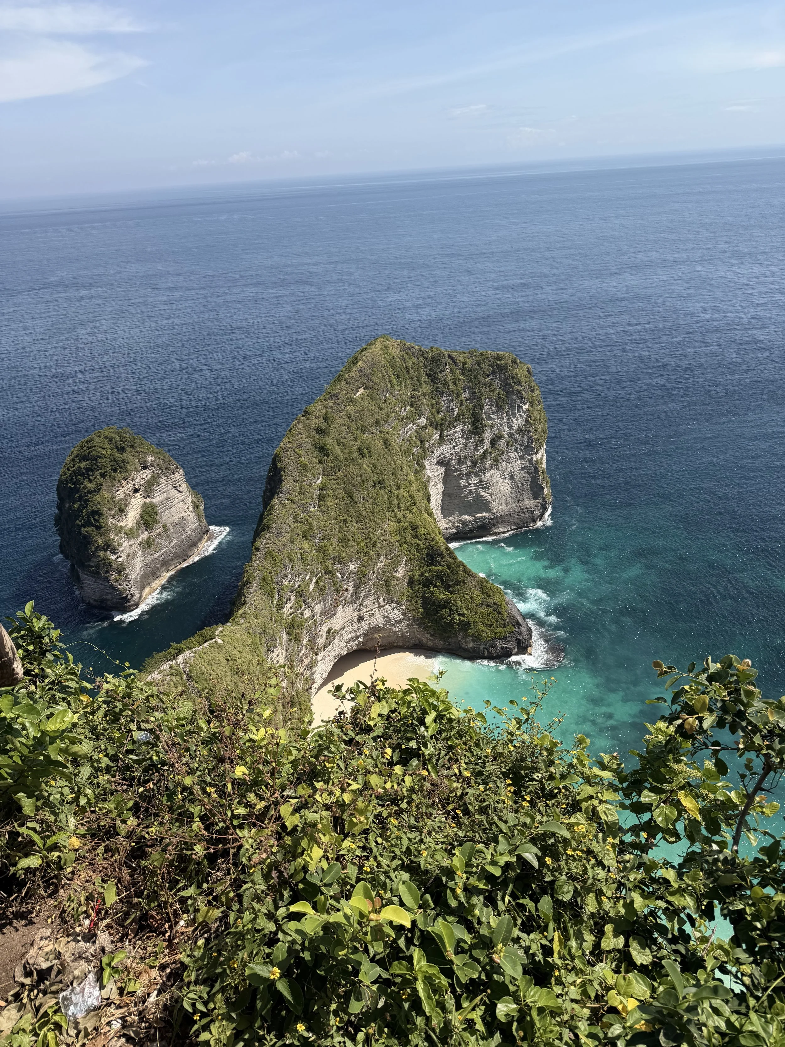 View of a rocky island with lush green vegetation, surrounded by turquoise water and a sandy beach at the base, with the ocean extending to the horizon under a blue sky.