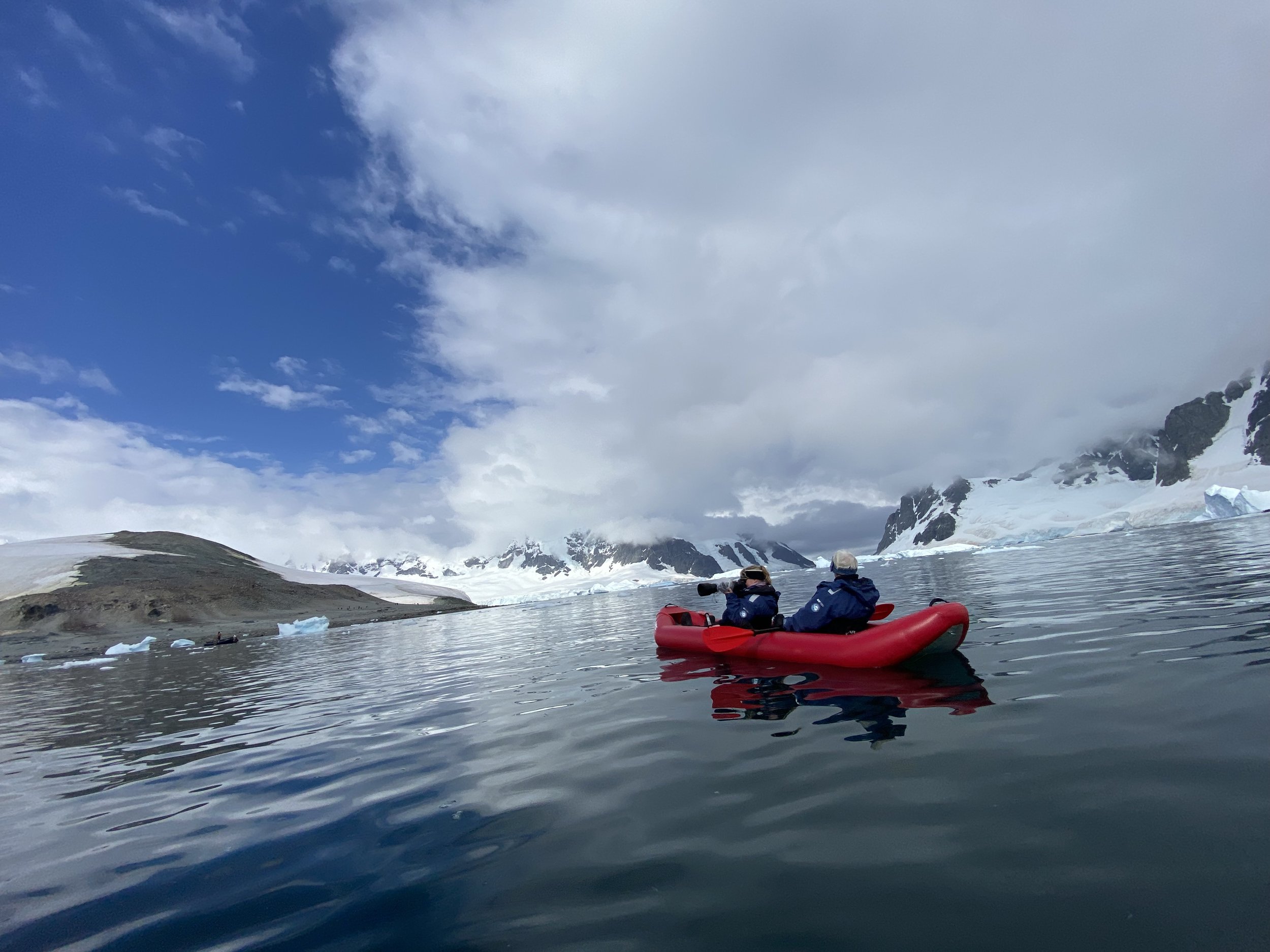 People in an orange kayak paddling on icy water in a snowy mountain landscape with cloudy sky.