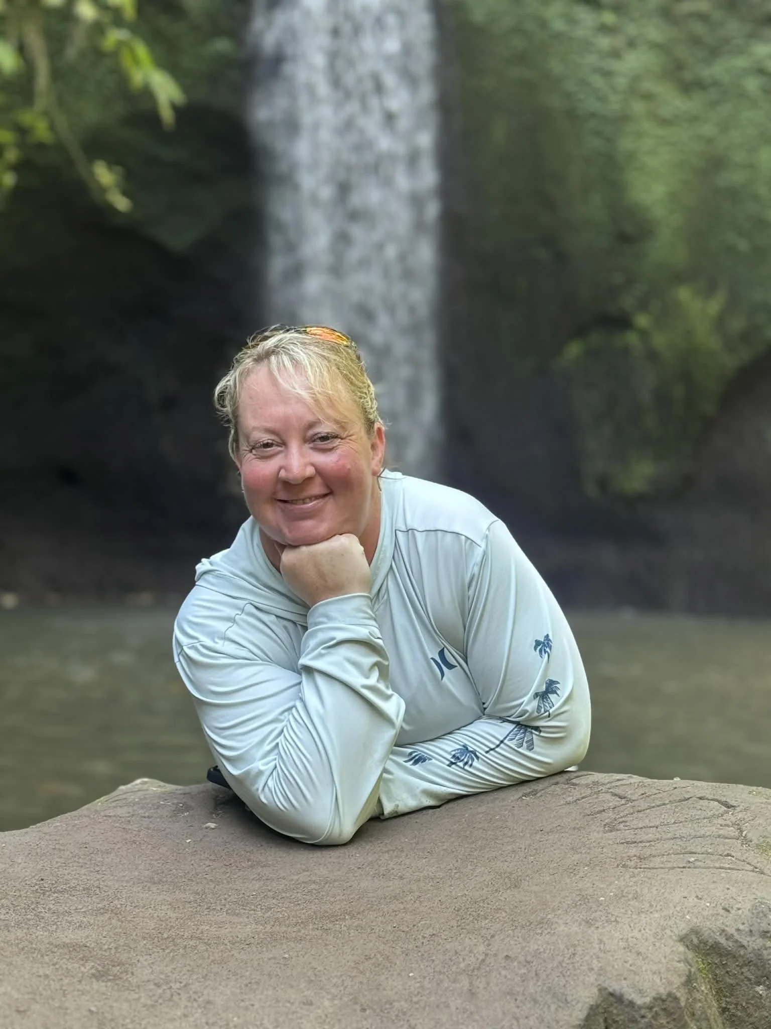Smiling woman with short blonde hair in a white long-sleeve shirt with palm tree pattern, lying on a large rock in front of a waterfall surrounded by greenery.