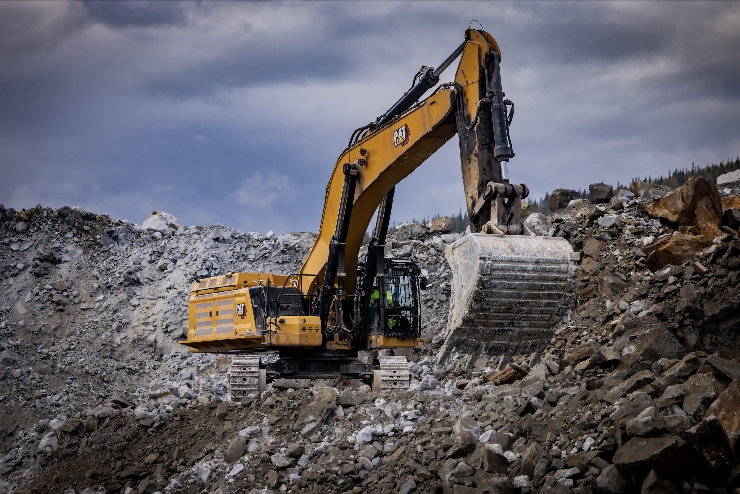 A yellow excavator working on a construction site with a pile of dirt, surrounded by a black barrier and a background of tall green trees and a blue sky.