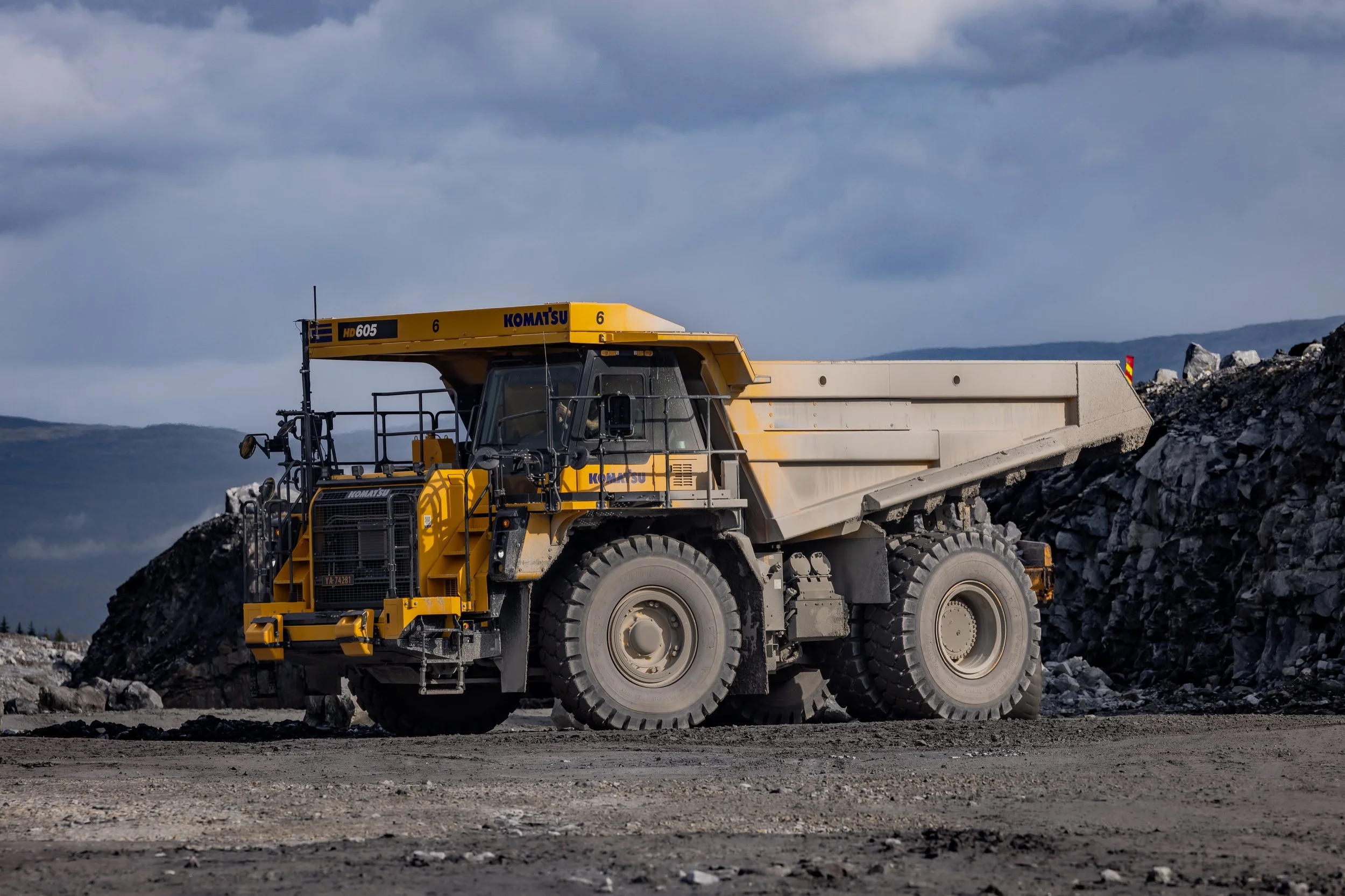 A large yellow Volvo articulated dump truck loaded with gravel, moving on a dirt road during a construction site near a body of water.
