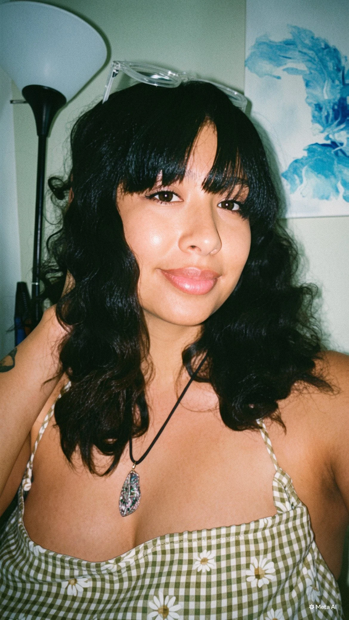A woman with curly black hair and a fringe, wearing a green and white checkered dress with daisies, sunglasses on her head, and a pendant necklace, smiling at the camera indoors with a green wall, a photo of water, and a black lamp in the background.