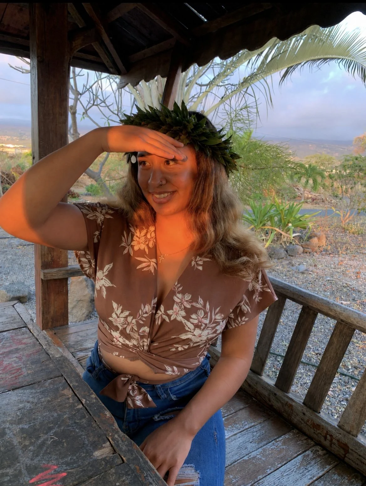 A woman with long wavy hair wearing a floral brown blouse and jeans is sitting outdoors under a wooden structure. She is wearing a leafy crown and shading her eyes with her hand while smiling, with a scenic background of desert landscape, some green plants, and a cloudy sky at sunset.