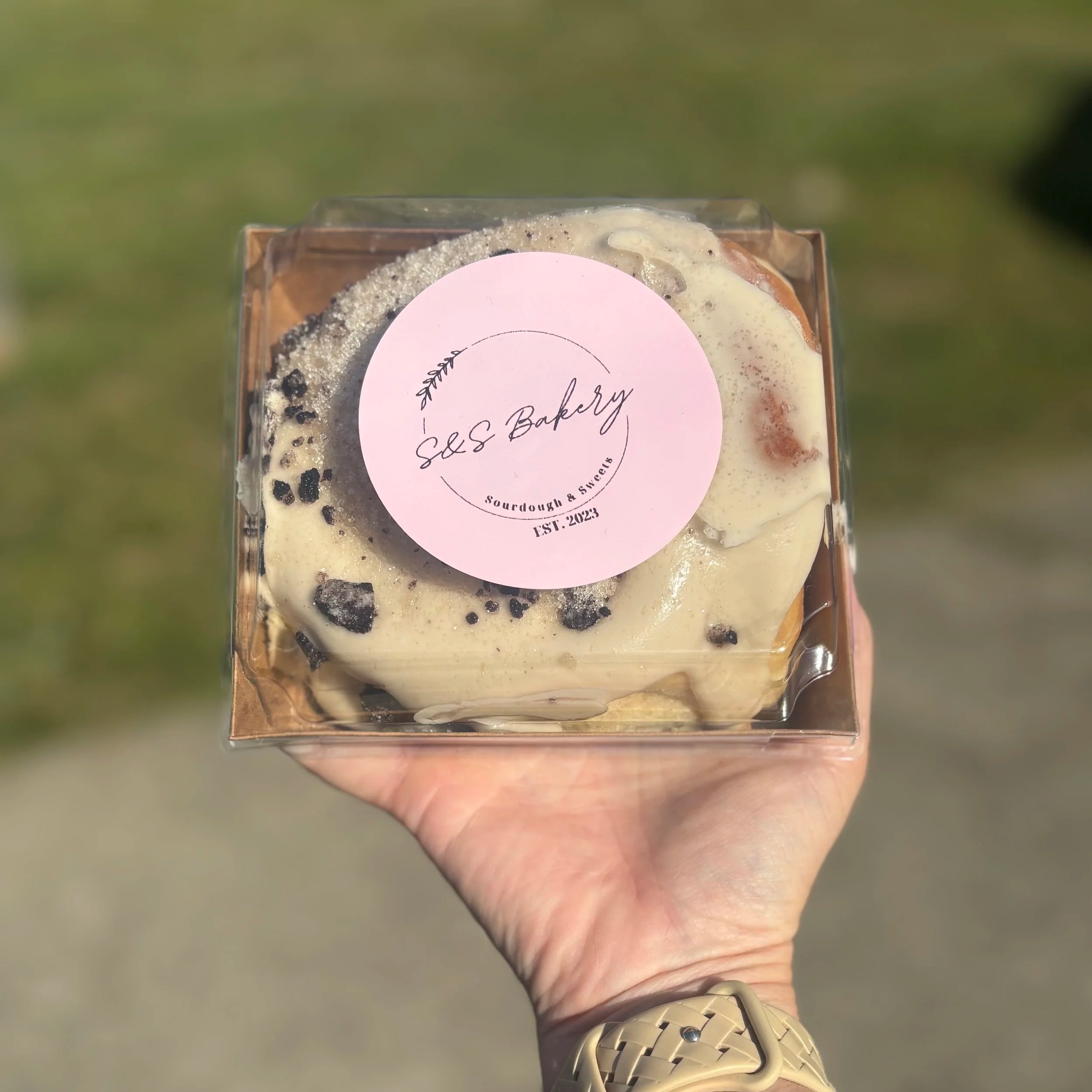 Close-up of a person's hand holding a bakery box with a pink circular label reading 'S&S Bakery, Sourdough & Sweets, Est. 2023'; the hand is wearing a beige watch, and the background is blurred green grass.