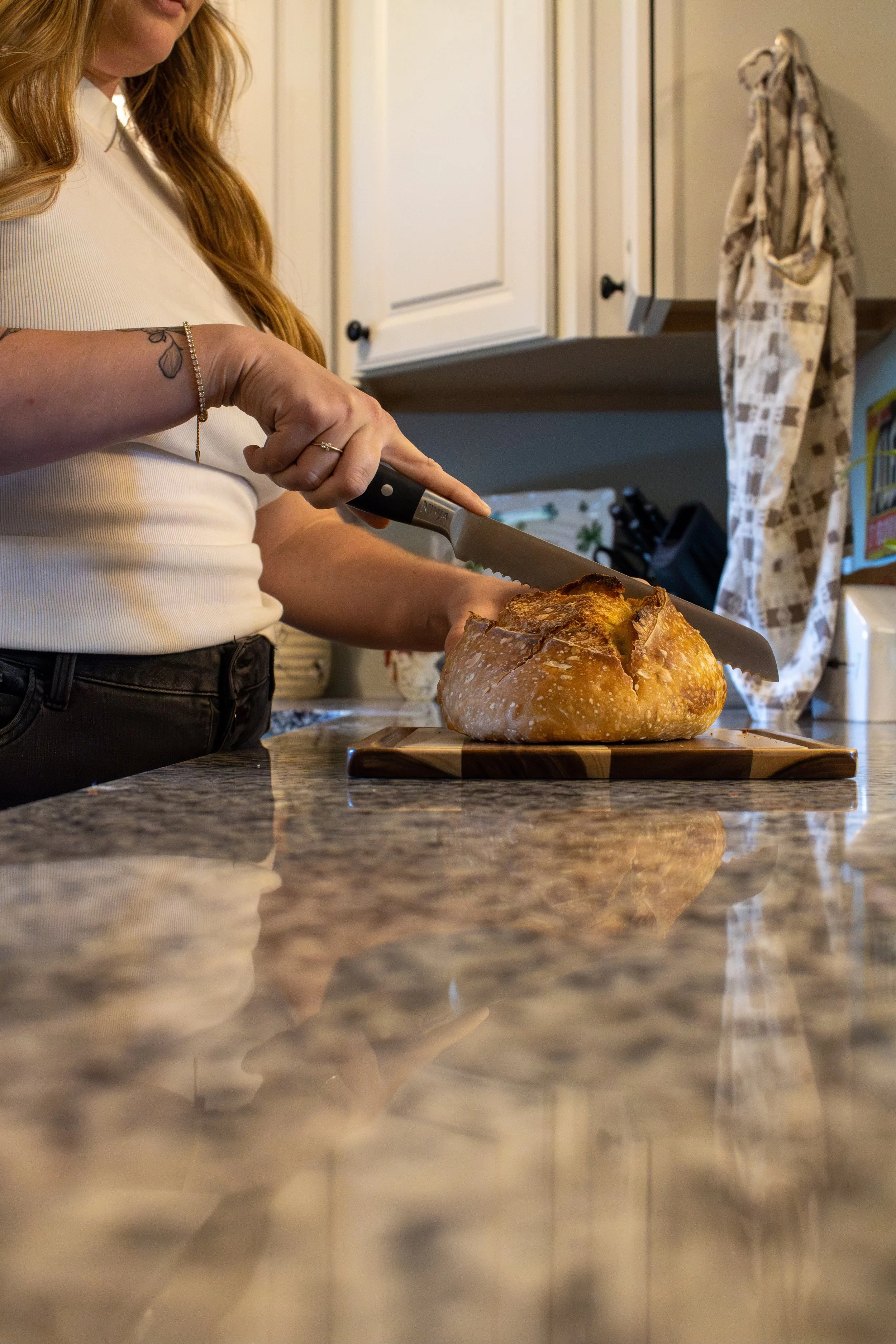 A woman with long red hair slicing a loaf of bread on a kitchen countertop with a large knife.