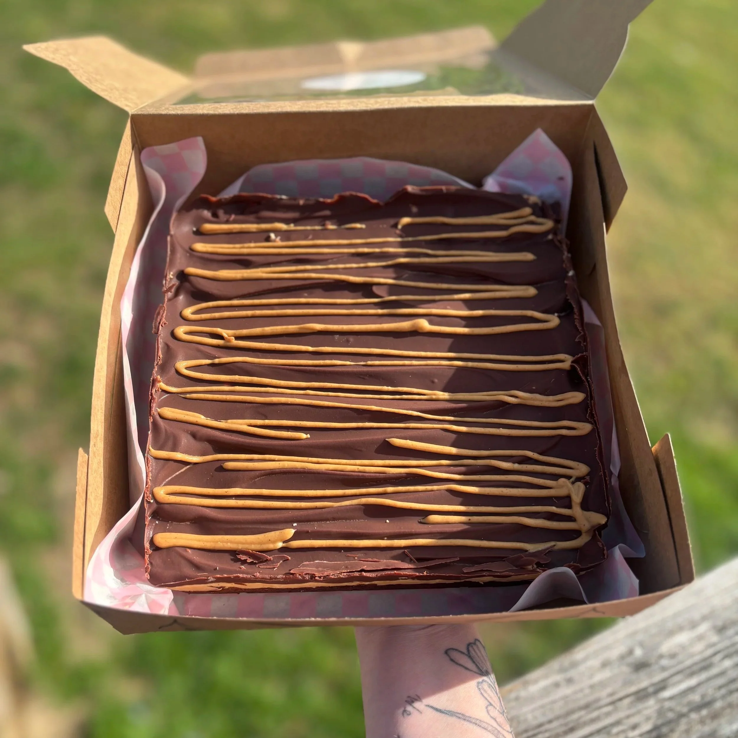 A person's hand holding an open box of chocolate cake with pink frosting and caramel drizzle, outdoors on a grassy area.