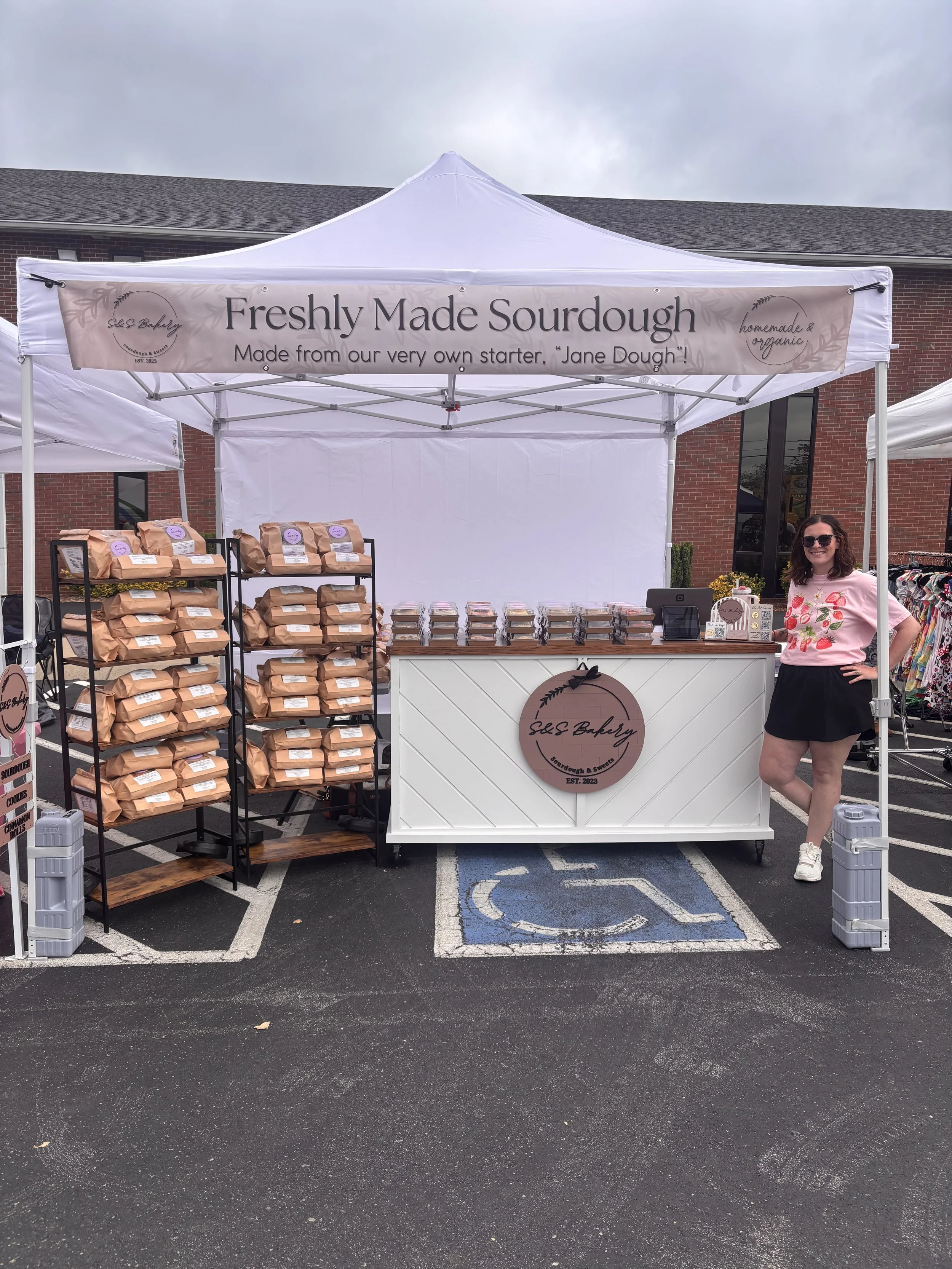 A woman standing next to a bakery stall with a sign that reads "Freshly Made Sourdough, Made from our very own starter, 'Jane Dough!'" The stall has baked sourdough bread in brown paper bags on racks and other baked goods displayed on the counter. The woman is wearing sunglasses, a pink shirt with strawberries, a black skirt, and white sneakers, and is smiling.