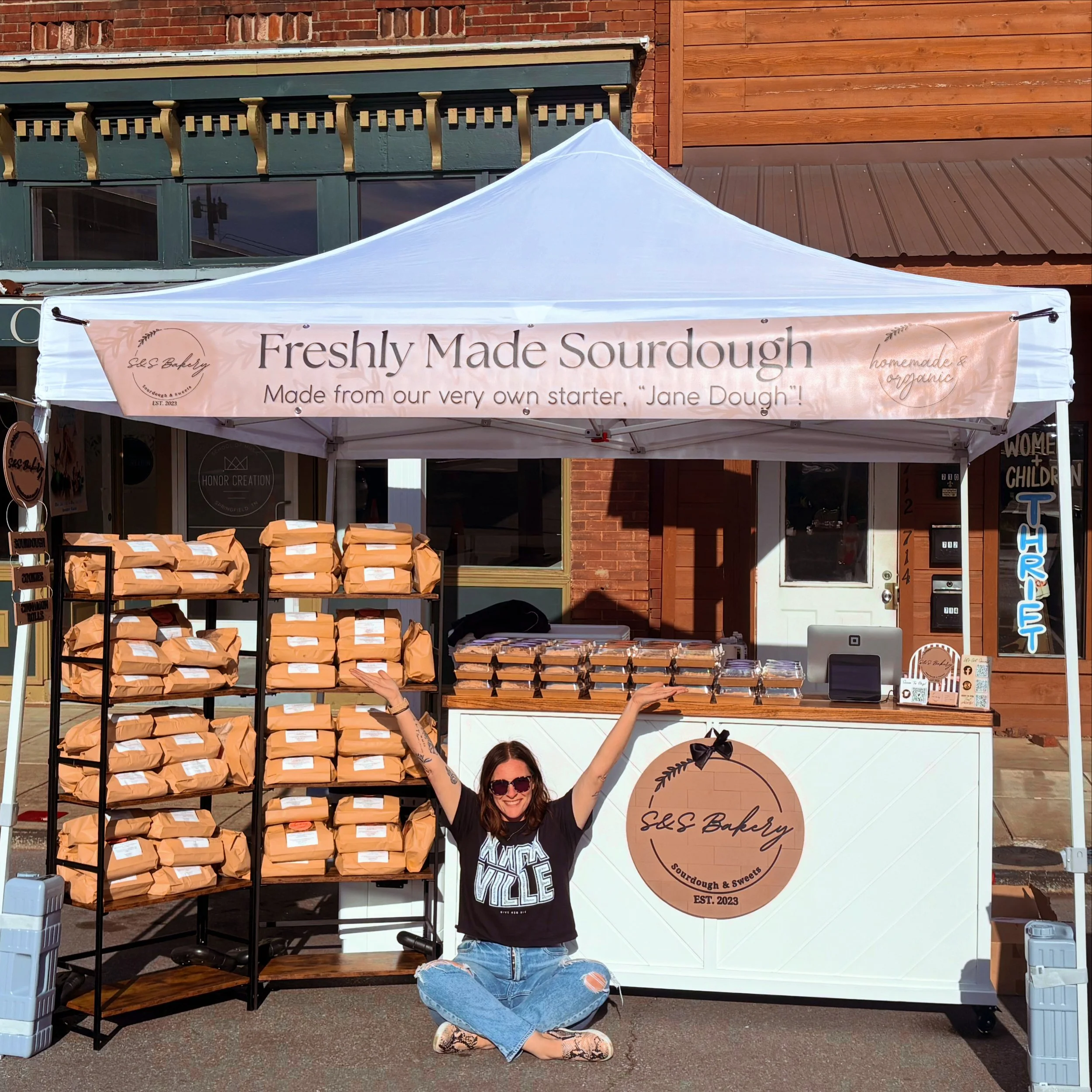 A woman with long hair and sunglasses is sitting on the ground, smiling, in front of a small bakery stand labeled 'Freshly Made Sourdough' with baked goods in paper bags. She is wearing a black t-shirt and ripped jeans, and she is raising her arm, pointing to the bakery sign.