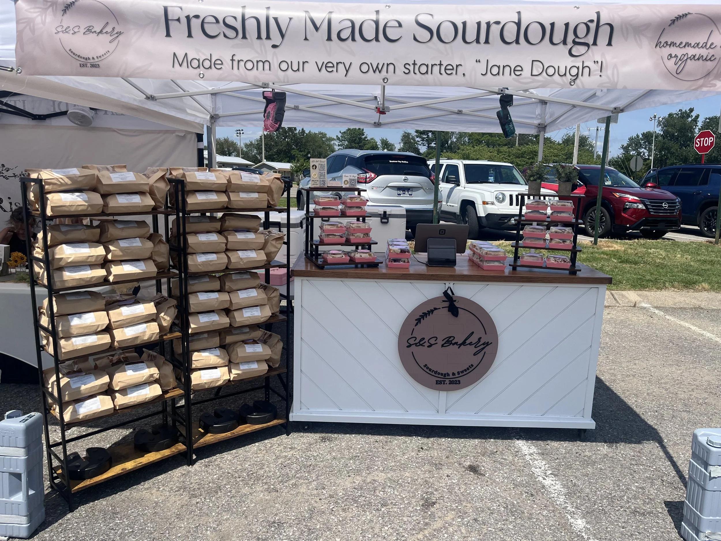 A market stall offering freshly made sourdough bread, with shelves of paper bags and prepackaged bread loaves. The stall is set up outdoors in a parking lot, with cars and a stop sign visible in the background.
