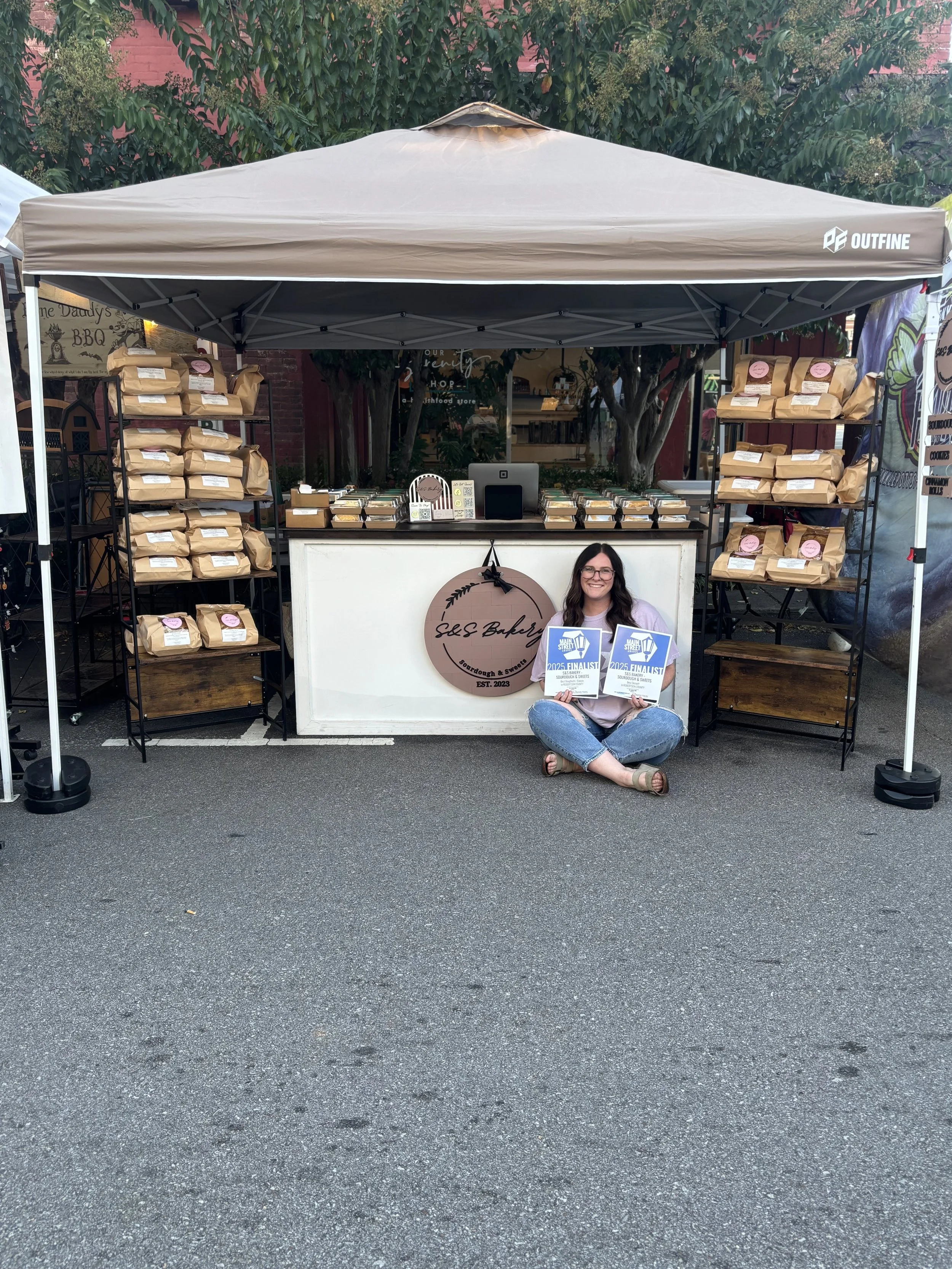 A woman sitting cross-legged on the ground in front of a stand, holding two signs that say '2025 Finalist'. The stand displays baked goods on shelves and has a sign that reads 'S&S Bakery'.