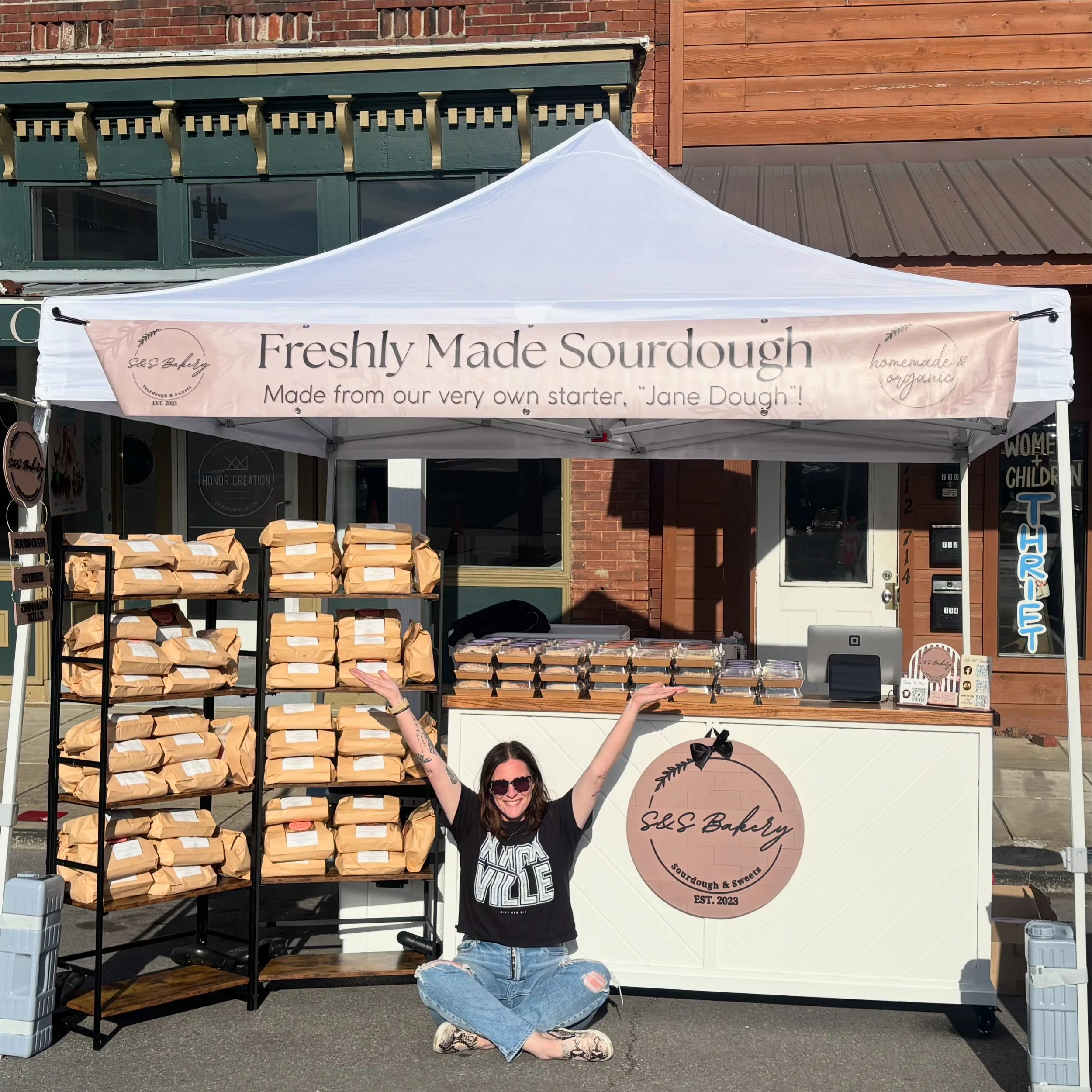 A woman with dark hair, wearing sunglasses, a black T-shirt, and ripped jeans, is sitting cross-legged in front of a white tent with a sign that reads "Freshly Made Sourdough" and "Sourdough & Sweets, Est. 2023." The tent display includes shelves with brown paper bags and containers of baked goods. The background shows a brick building with windows.