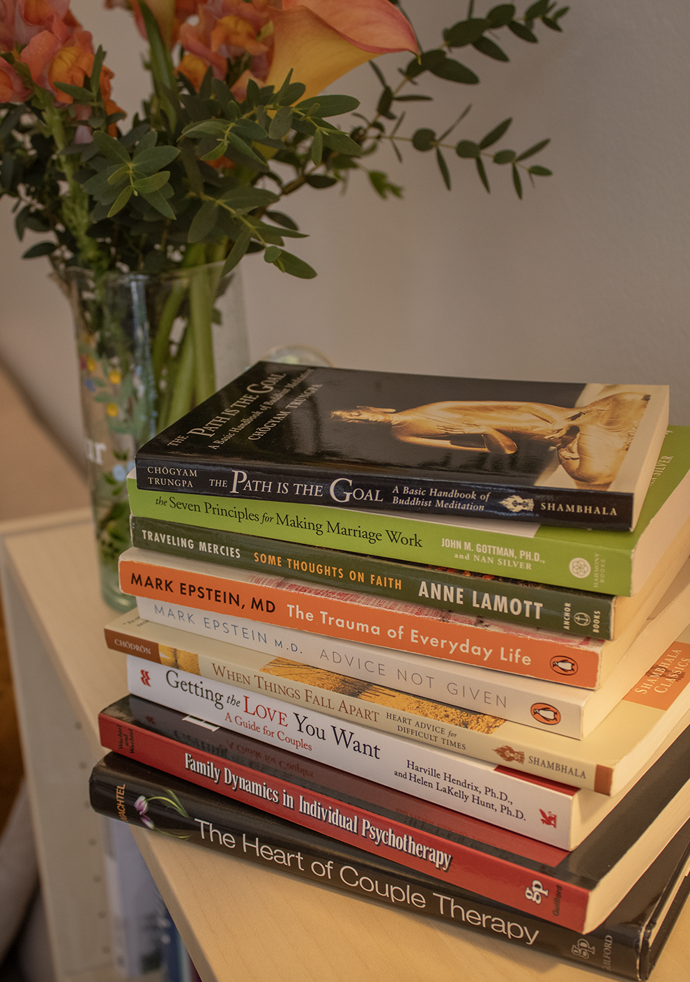 A stack of ten books on a small table, with a vase of multicolored flowers next to them.