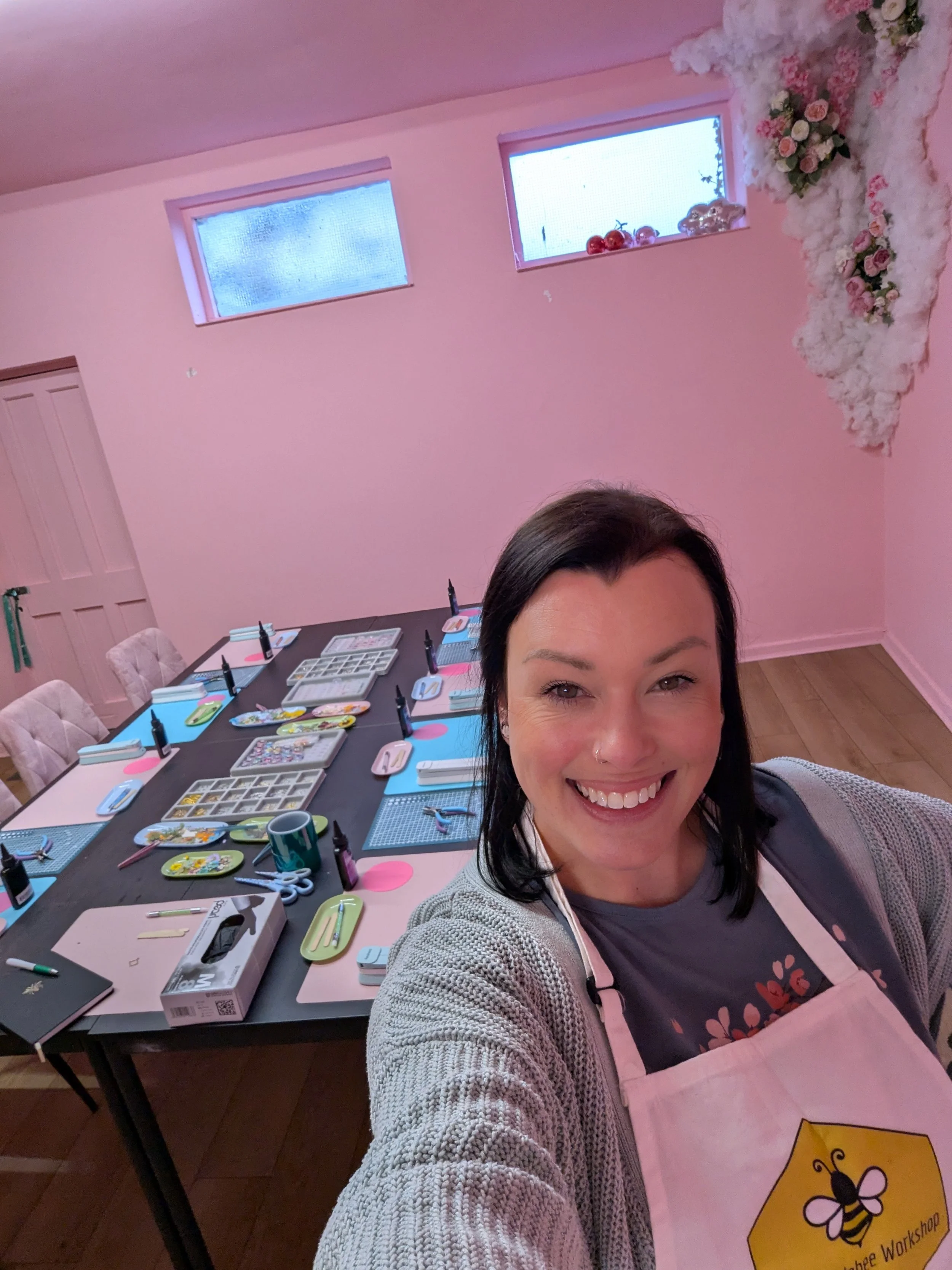 A woman smiling in a craft room with a large table covered in art supplies, pink walls, and flower decorations on the wall.