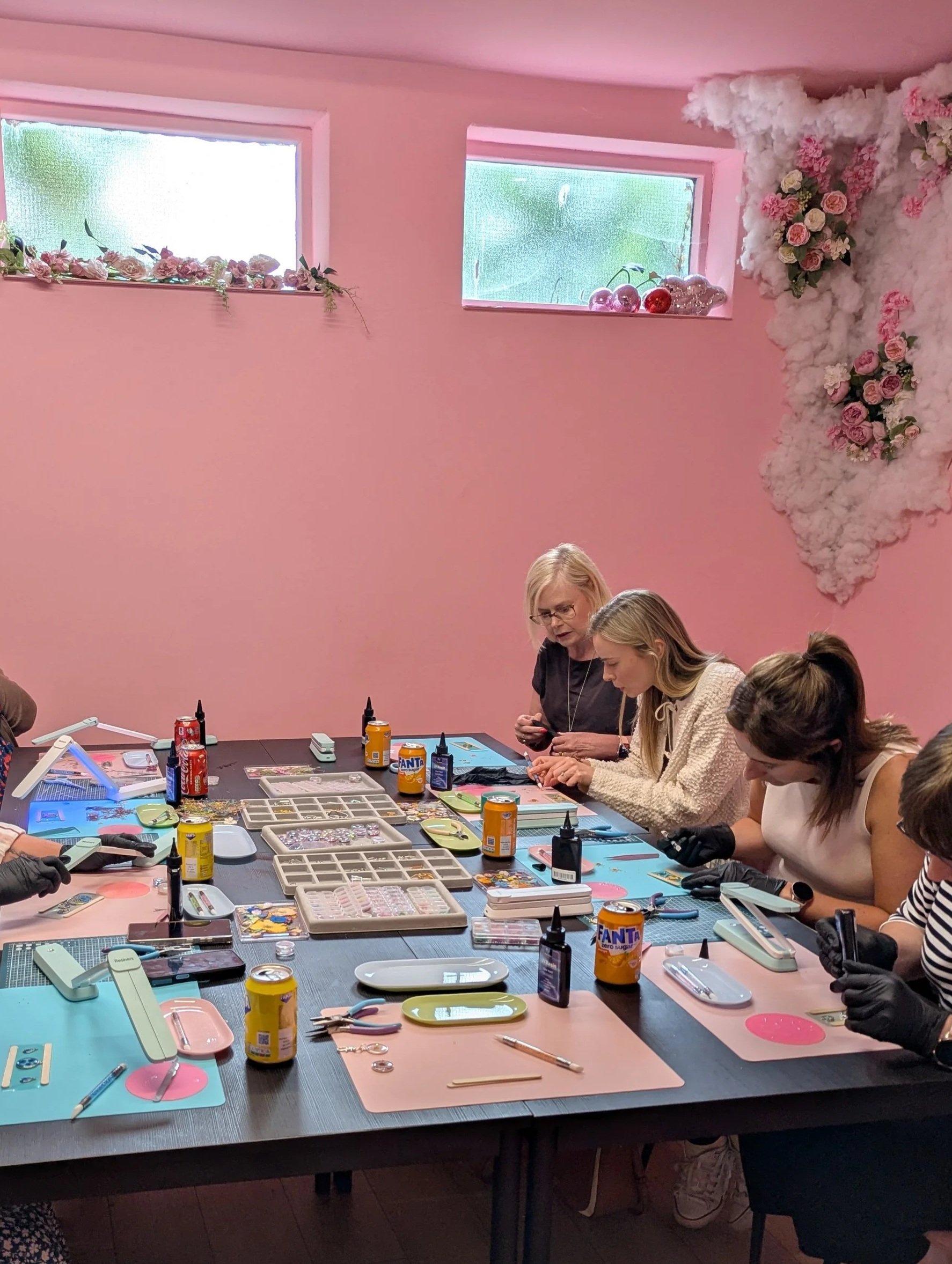 People sitting at a table engaged in a crafting activity, with various craft supplies, tools, and beverages on the table against a pink wall with flower and cloud decorations.