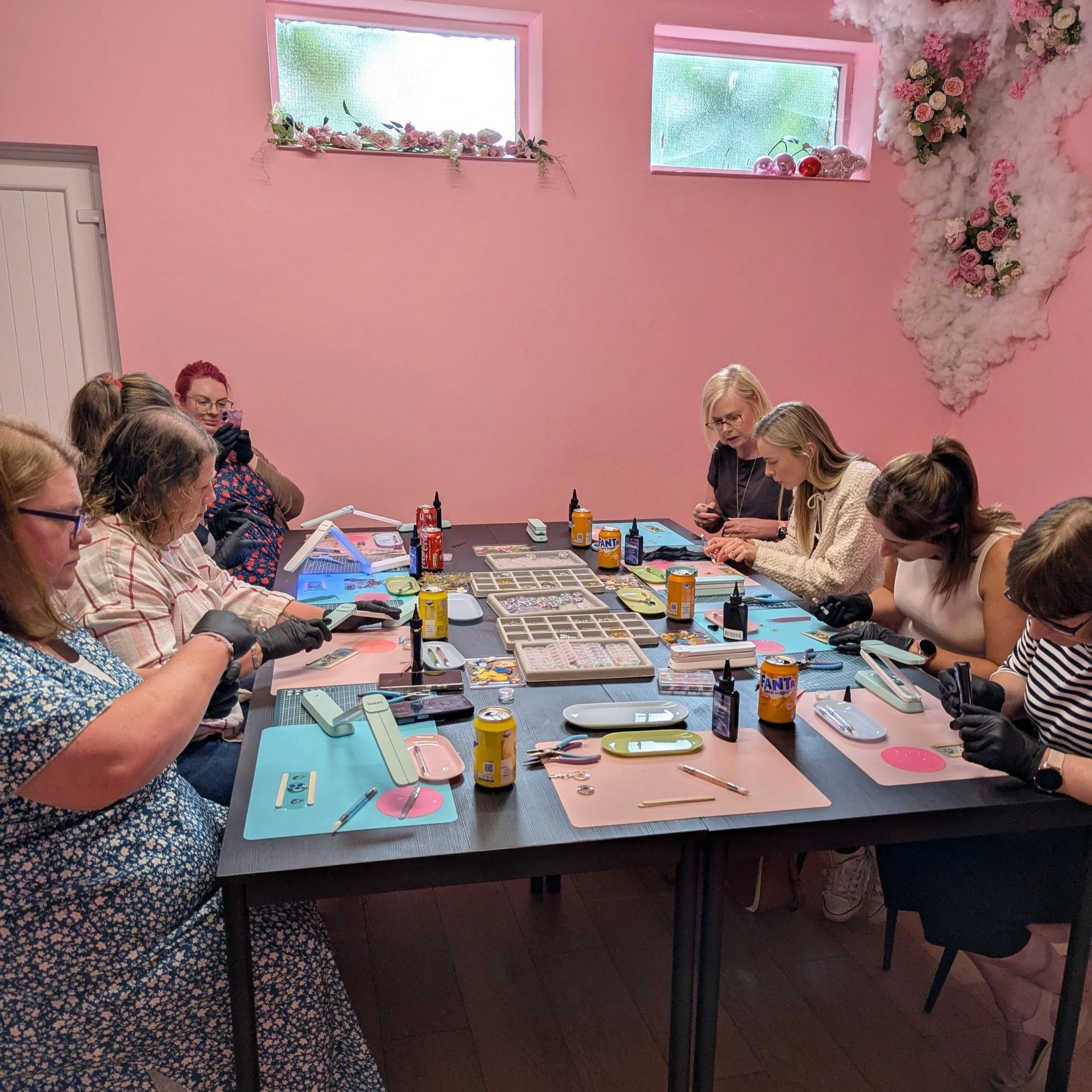 Group of women gathered around crafting table with supplies, bottles, and tools, in a pink-walled room decorated with floral arrangements and windows.