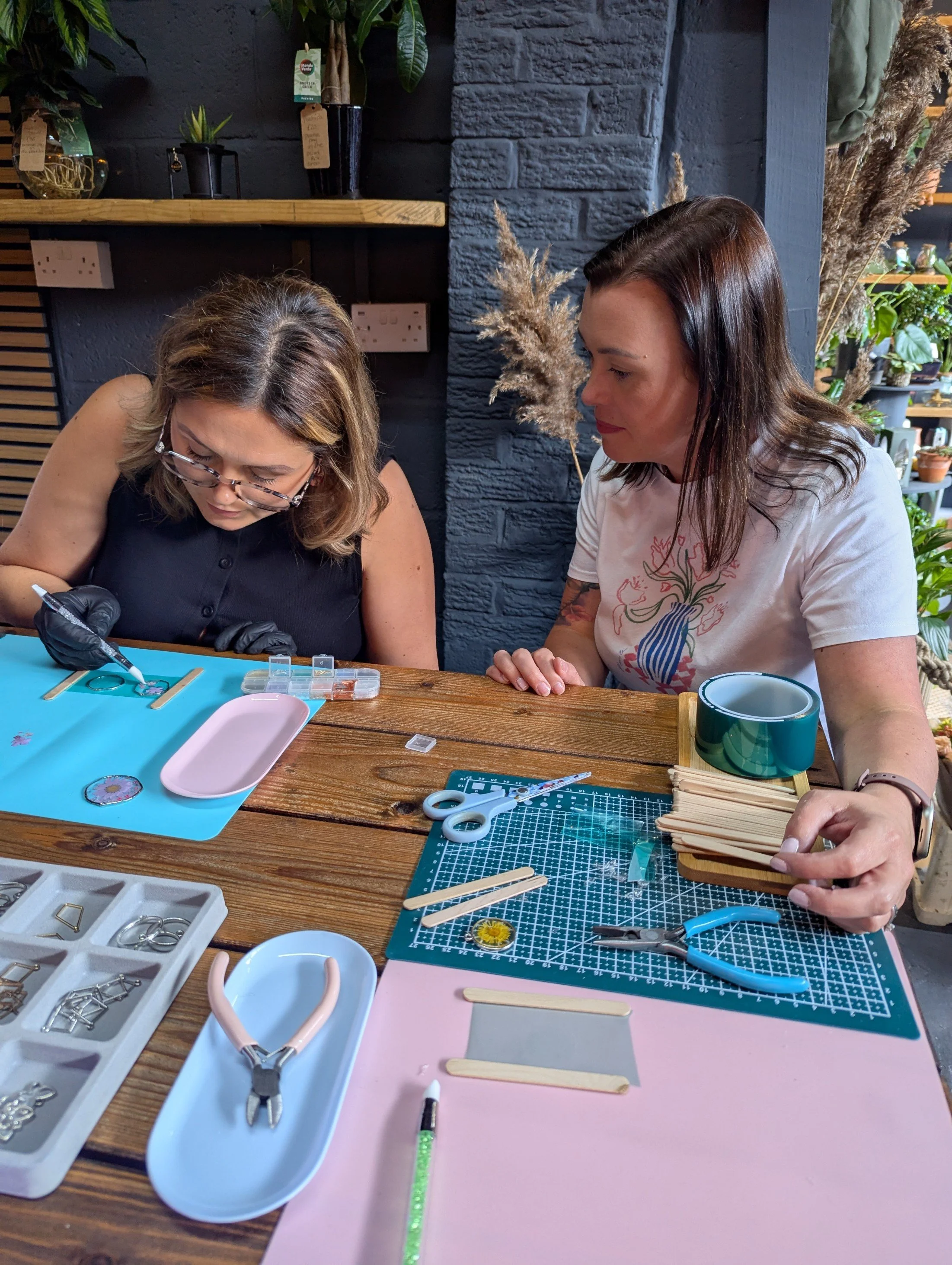 Two women are seated at a wooden table, engaged in a jewelry-making activity. The table is covered with various tools, jewelry findings, and crafted pieces. The background features potted plants on shelves and decorative dried grasses against a dark-colored brick wall.