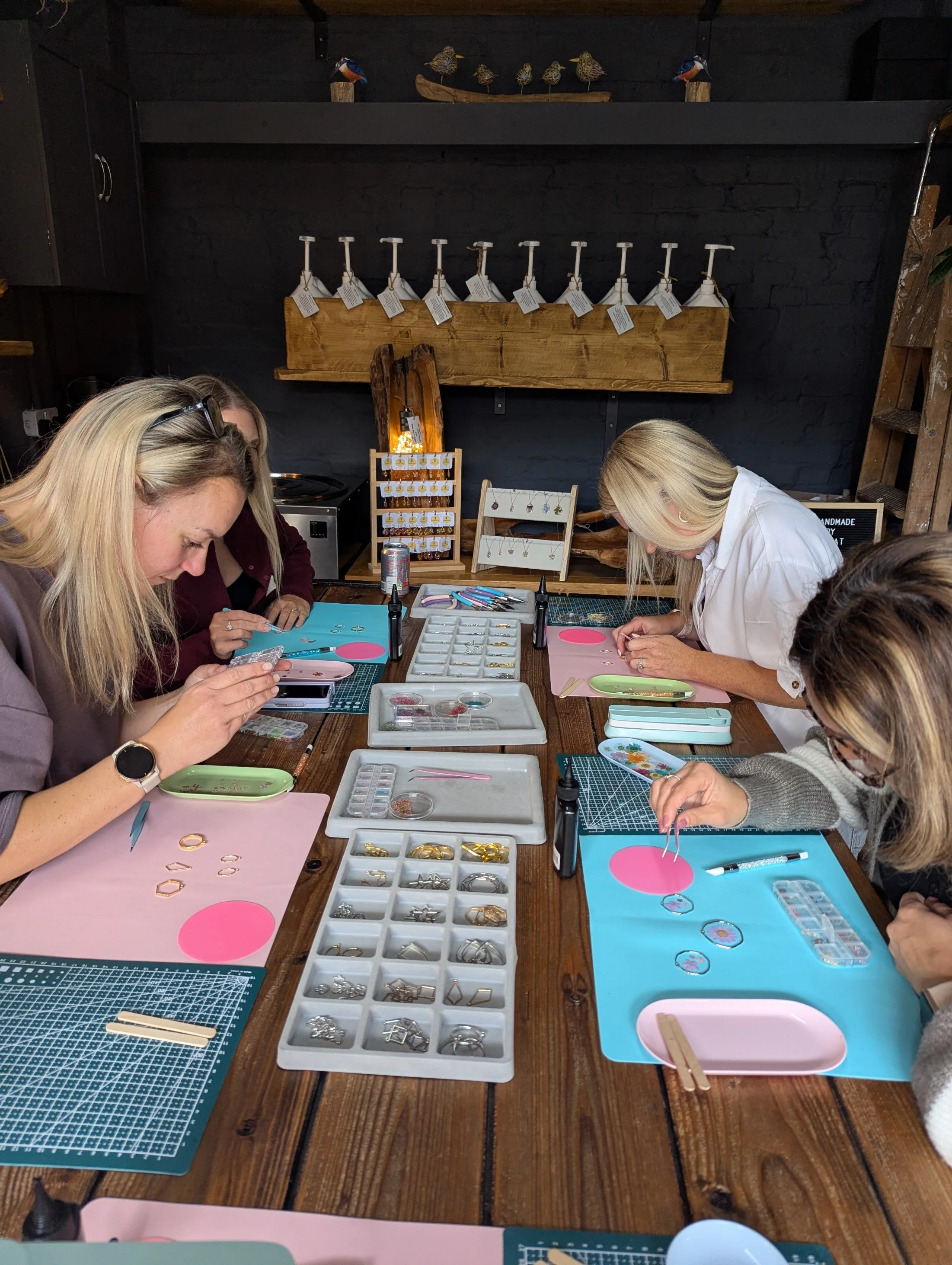 People sitting at a wooden table engaged in jewelry making with various tools and supplies, including trays of assorted jewelry pieces, colorful mats, and small containers, in a craft workshop setting.
