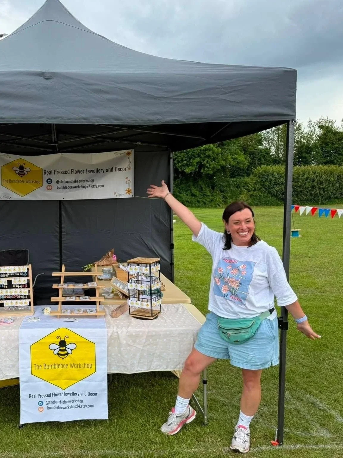 A woman standing next to a booth at an outdoor event, smiling and pointing to her right. The booth is under a gray canopy and displays jewelry and decorations, with a sign that reads 'The Bumblebee Workshop' and mentions real pressed flower jewelry and decor.