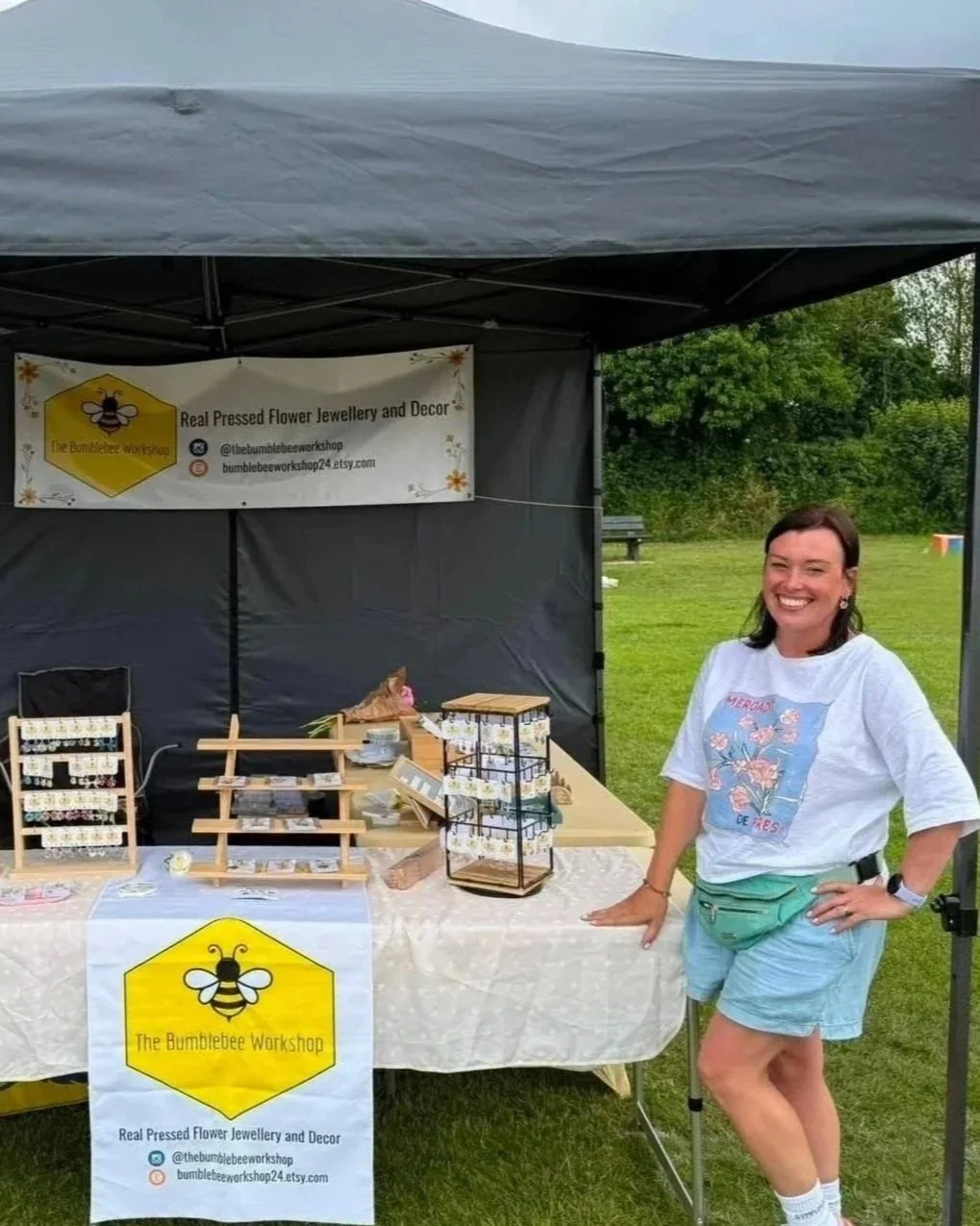 Smiling woman standing next to a booth selling pressed flower jewelry and decor, with a sign reading 'The Bumblebee Workshop' at an outdoor event.