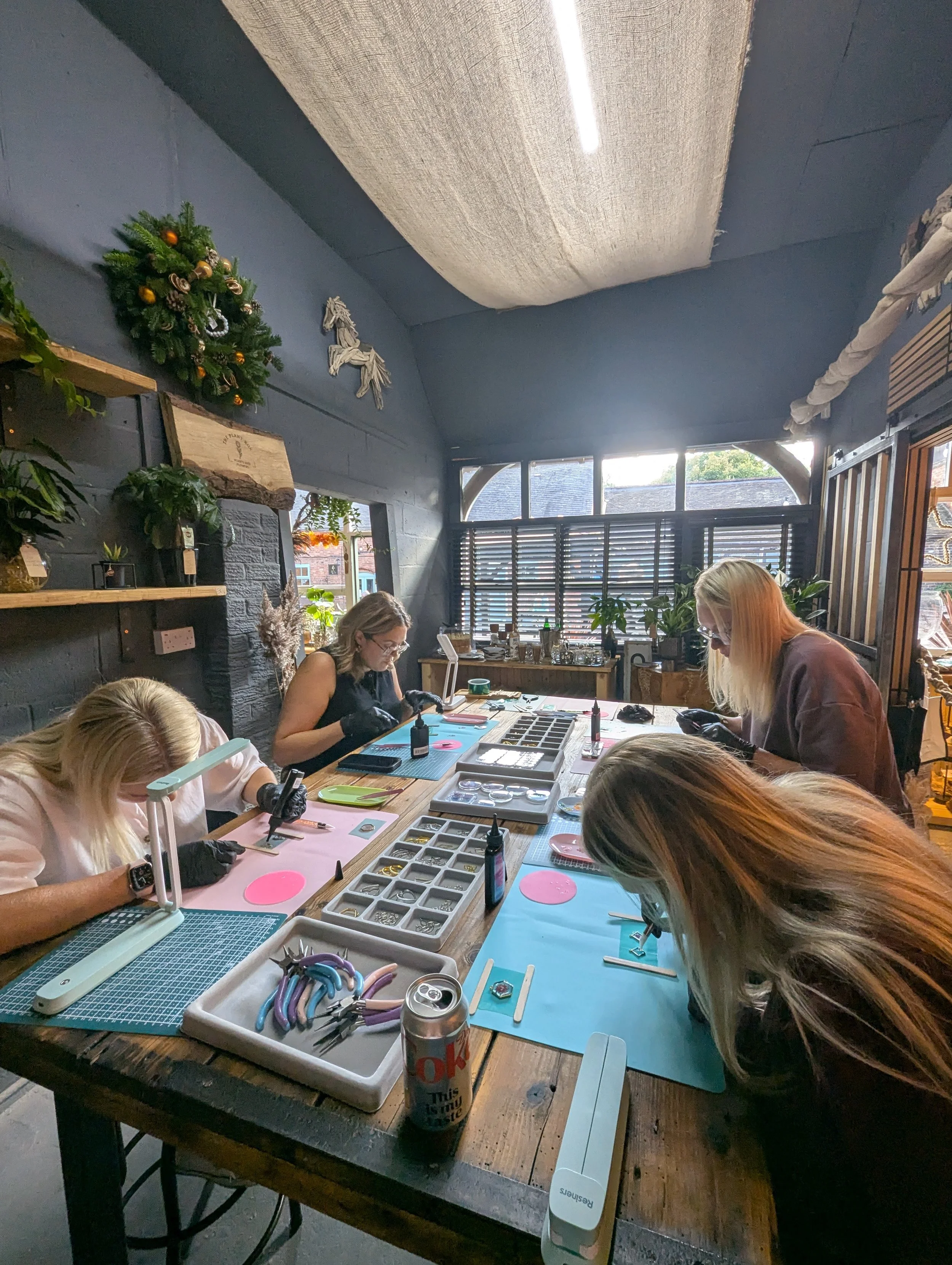 People working on jewelry or small crafts at a large wooden table in a well-lit room with large windows and decorative wall art, including a green holiday wreath.