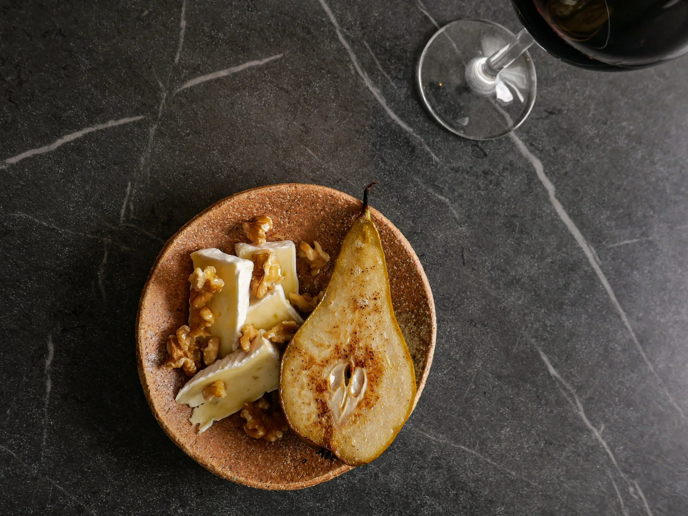 A round, earthy-colored ceramic plate on a dark, textured surface holds a wedge of cheese with walnuts, a slice of pear, and another piece of cheese topped with walnuts. A glass of red wine is partially visible in the top right corner.