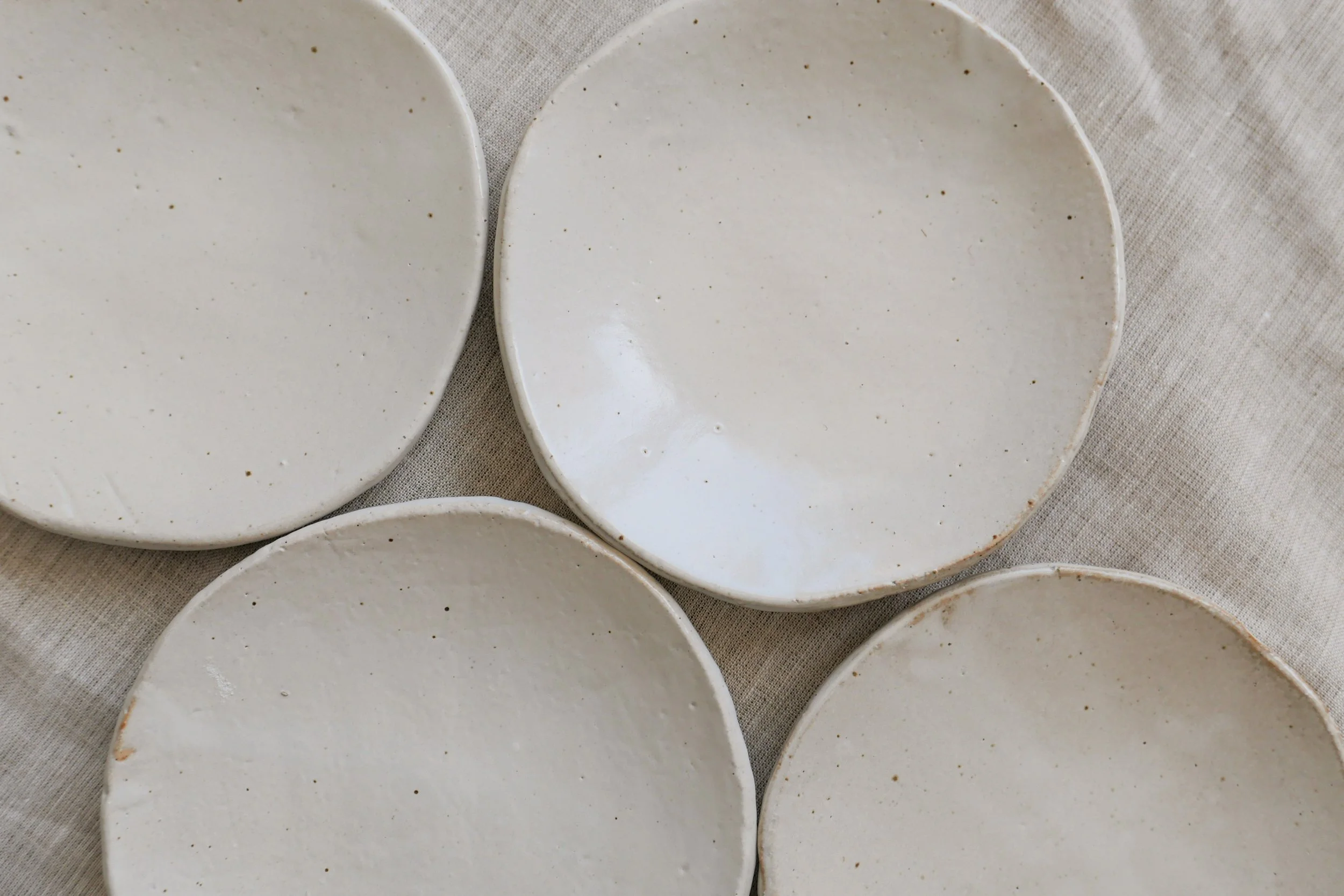 four titanium white plates by shigaraki seinenryo, seen from above on a linen cloth, showing the warm, milky white glaze with subtle dark speckles and gently irregular rims.