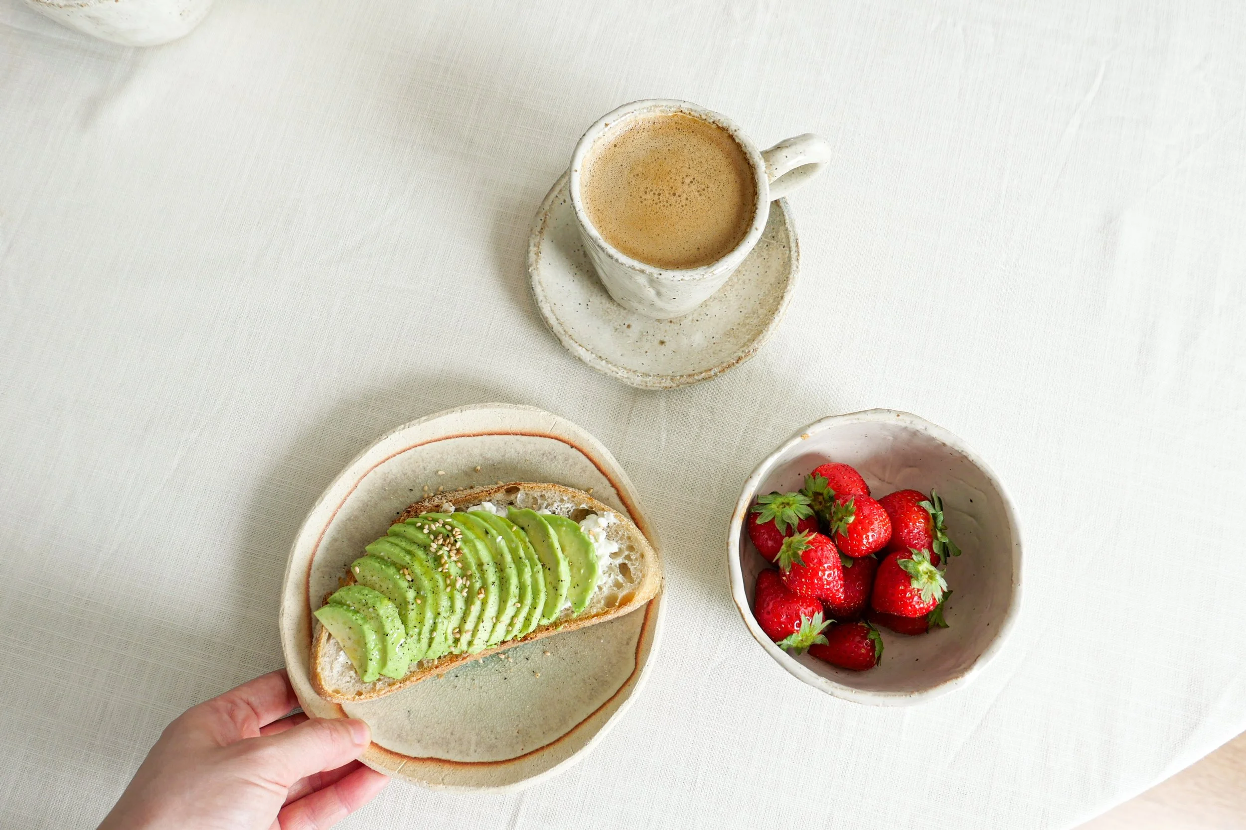A hand holding a toast with sliced avocado on top, surrounded by a bowl of strawberries and a cup of coffee on a white table.