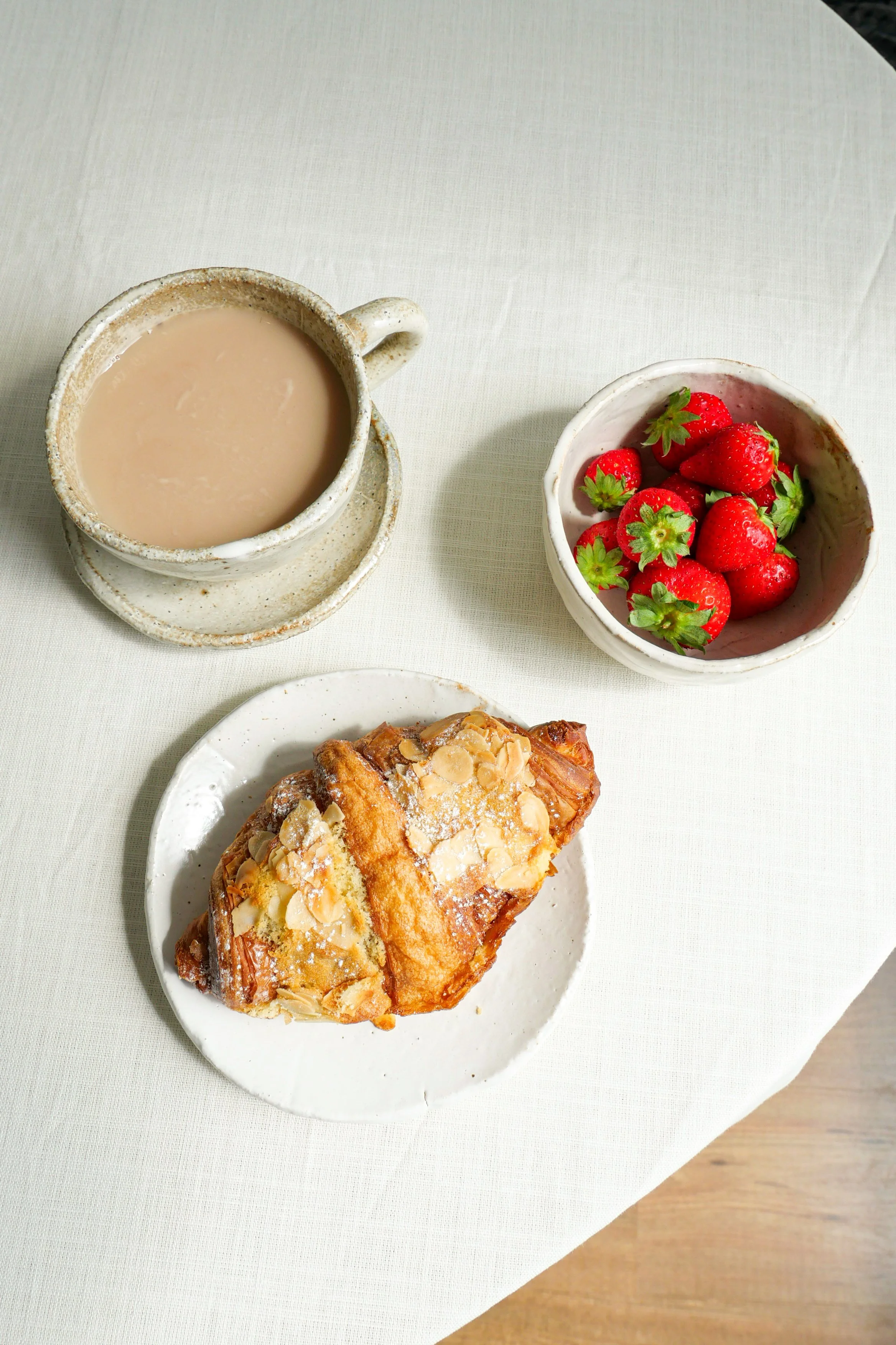 A ceramic mug of milky coffee, a bowl of fresh strawberries with green leaves, and a pastry with almond slices on a white plate, on a white tablecloth.