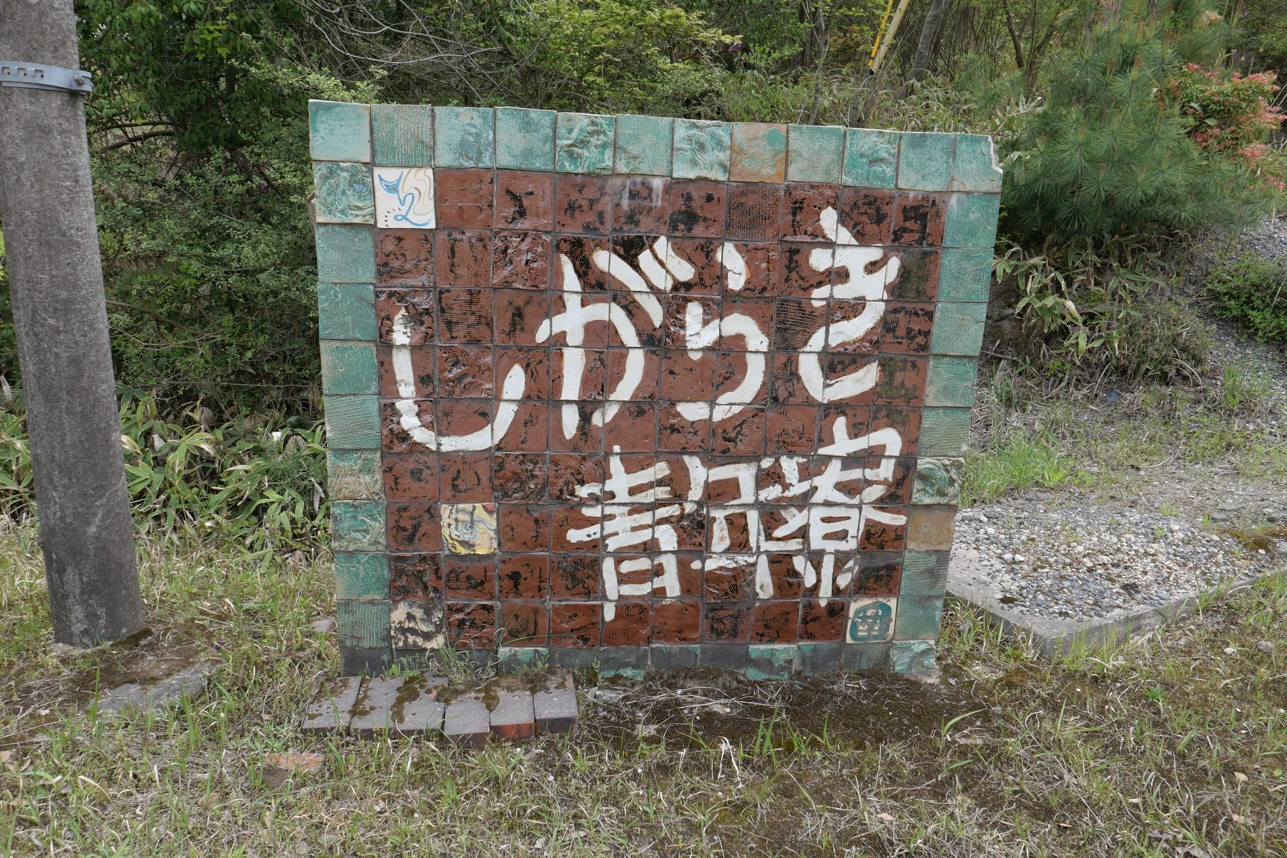 A ceramic tile sign reading "Shiragaki Seinenryo" set among grass and trees, made from Shigaraki ware tiles in earthy red and green
