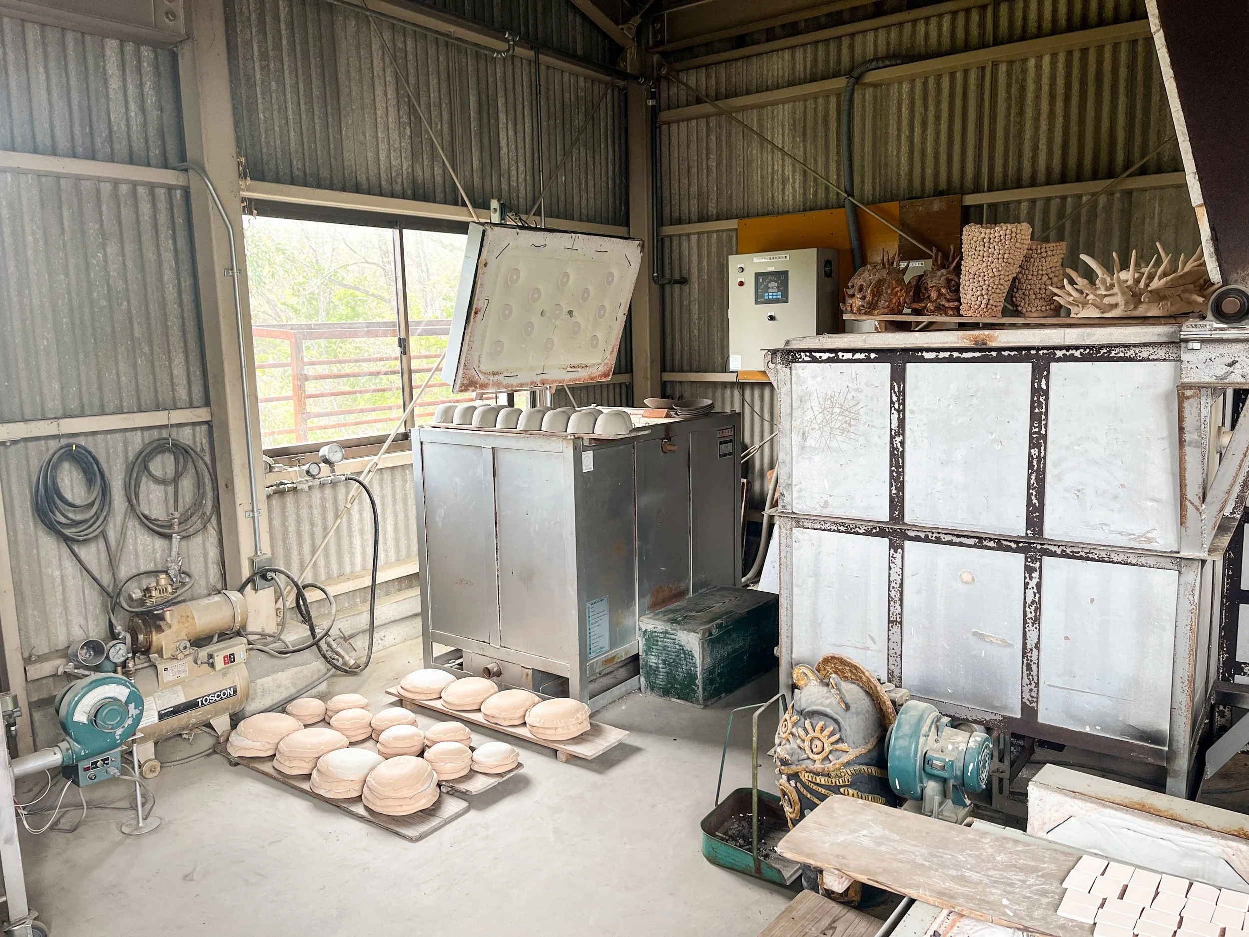 Artist's studio with clay sculptures of animal heads on a shelf, clay plates on a table, and various equipment for pottery work, with metal walls and a window letting in natural light.