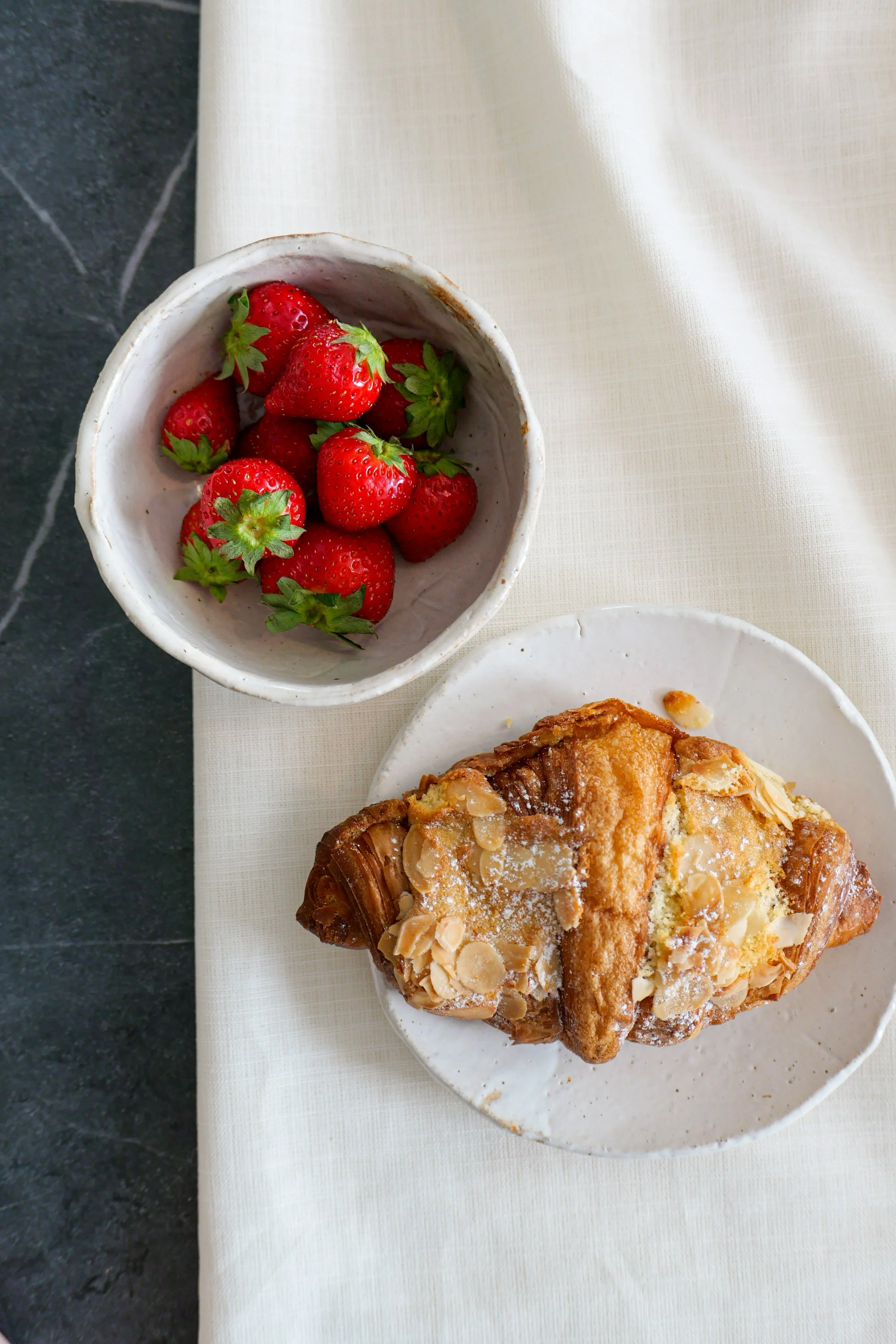 A bowl of fresh strawberries on a white tablecloth, with part of a pastry topped with sliced almonds visible below.