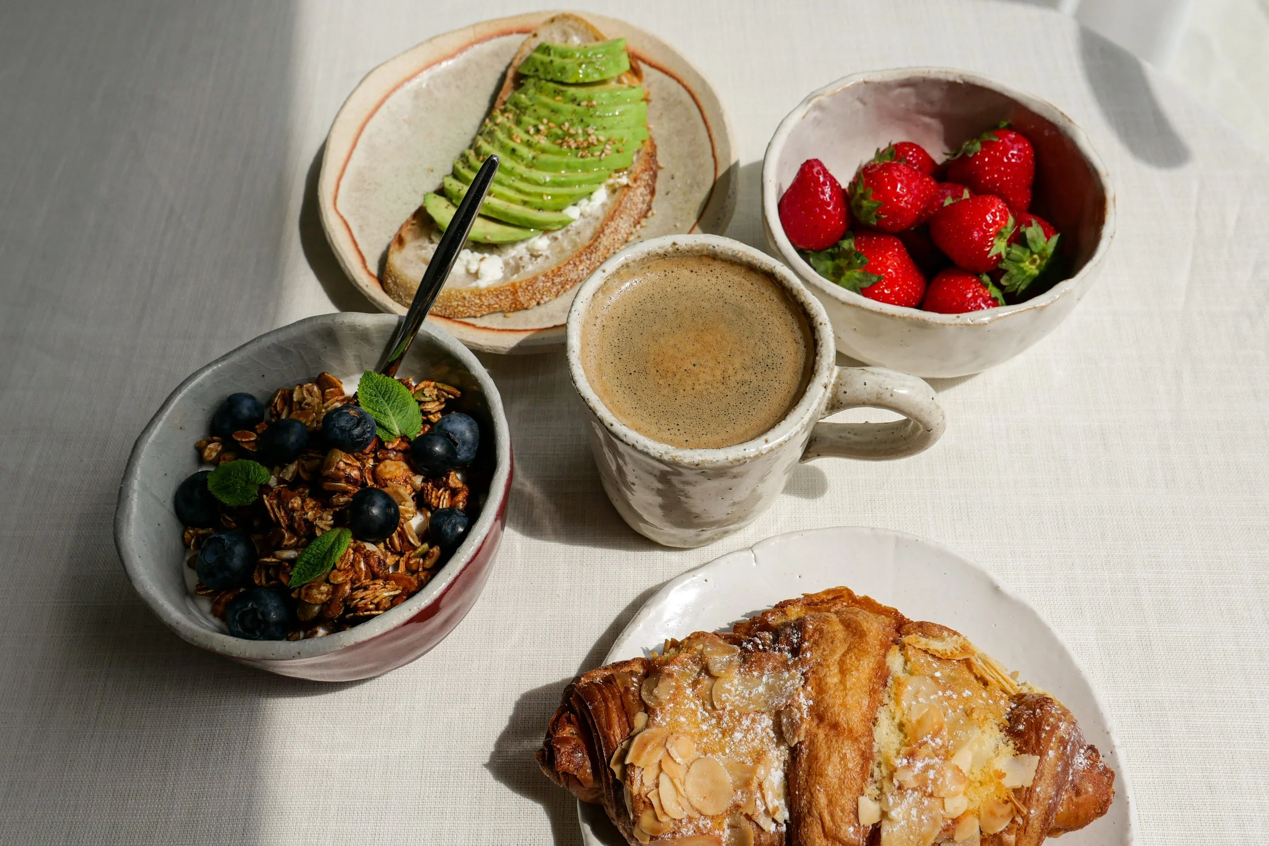 Assorted breakfast foods on plates, including strawberries, blueberries with granola, avocado toast with sesame seeds, coffee, and a pastry with almonds.