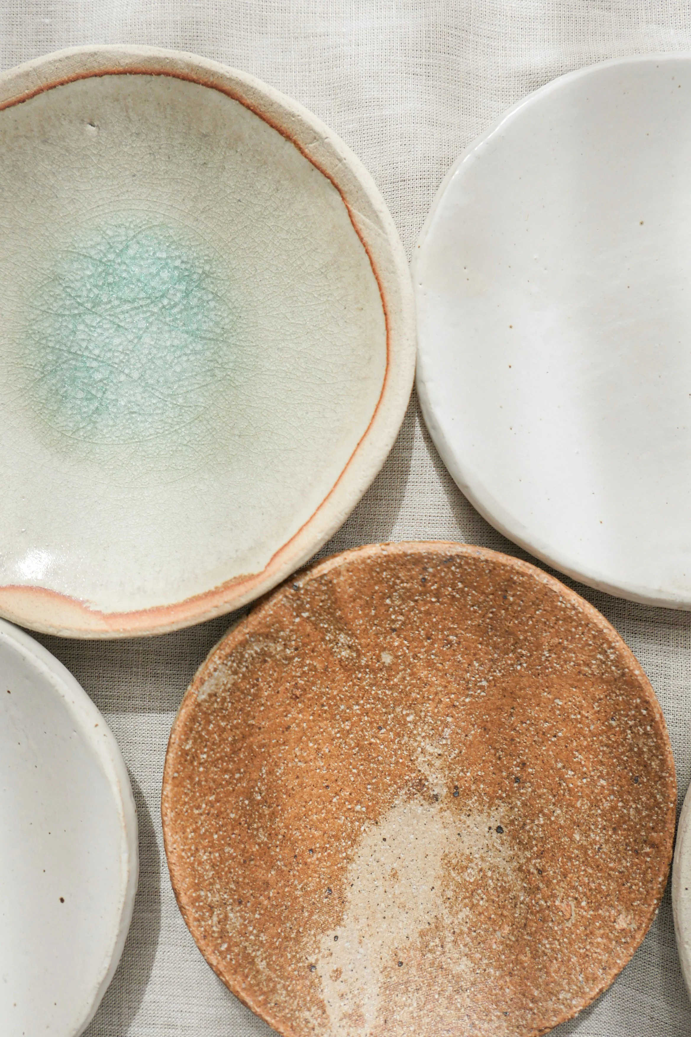 Close-up of various ceramic plates, including a white plate with a green glaze, another white plate with small speckles, and a brown speckled plate, all on a light-colored fabric surface.
