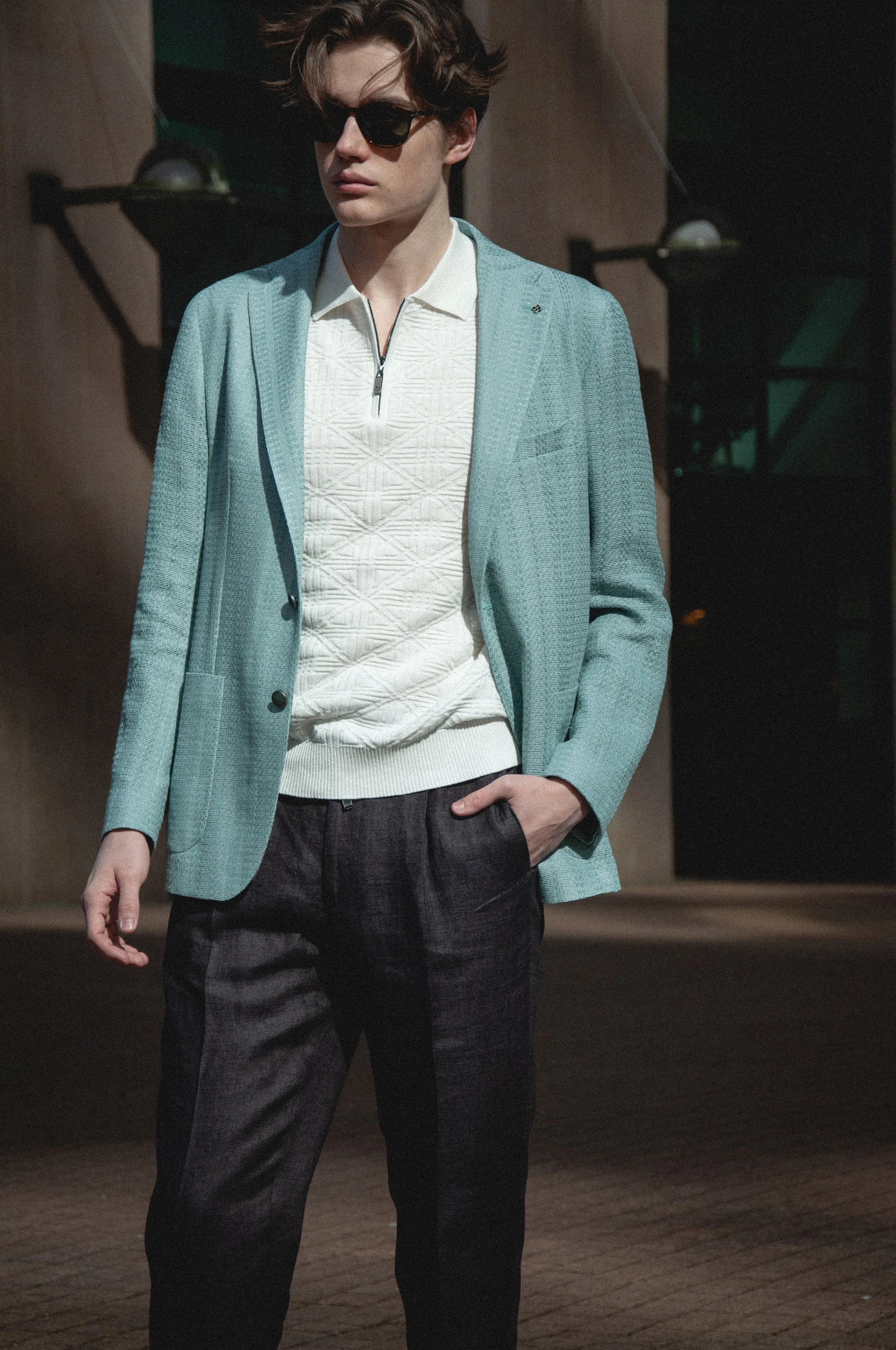 A man wearing black sunglasses, a white zipped shirt, a light blue blazer, and dark pants, standing outdoors with a brown building in the background.