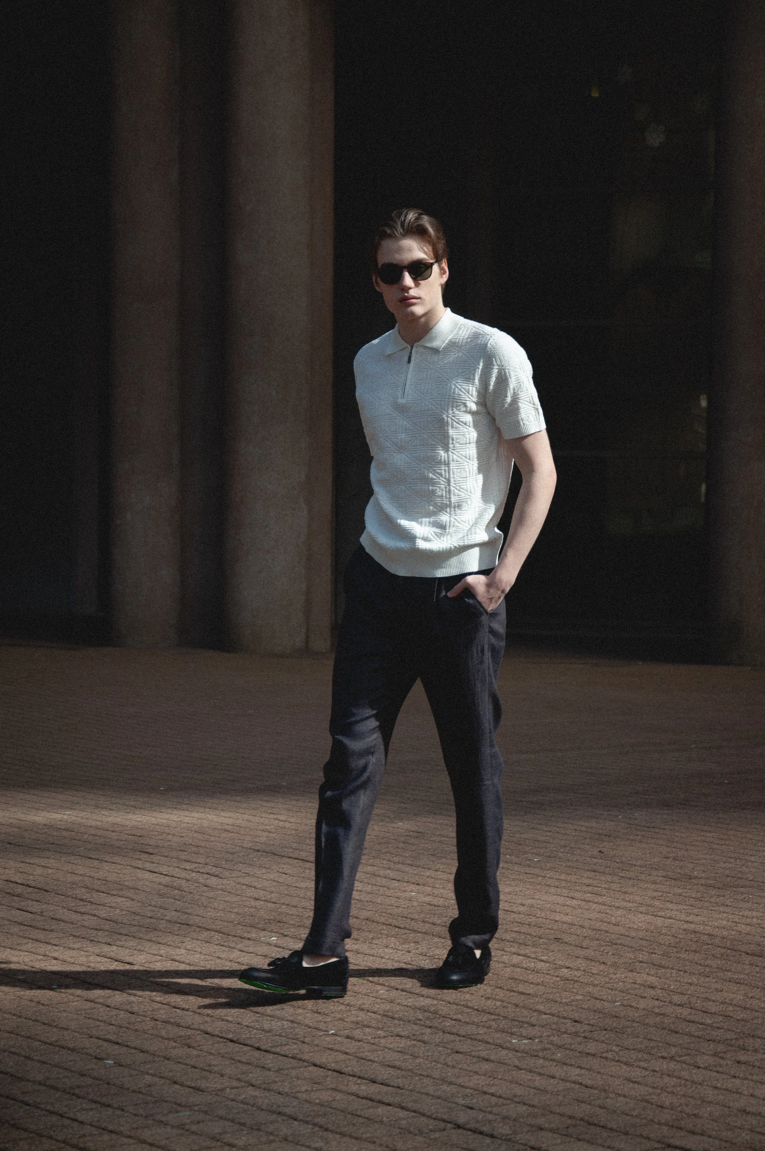 A young man with light skin, wearing sunglasses, a white textured polo shirt, dark pants, and black shoes, standing outdoors on a brick-paved surface, with a dark background and large columns.