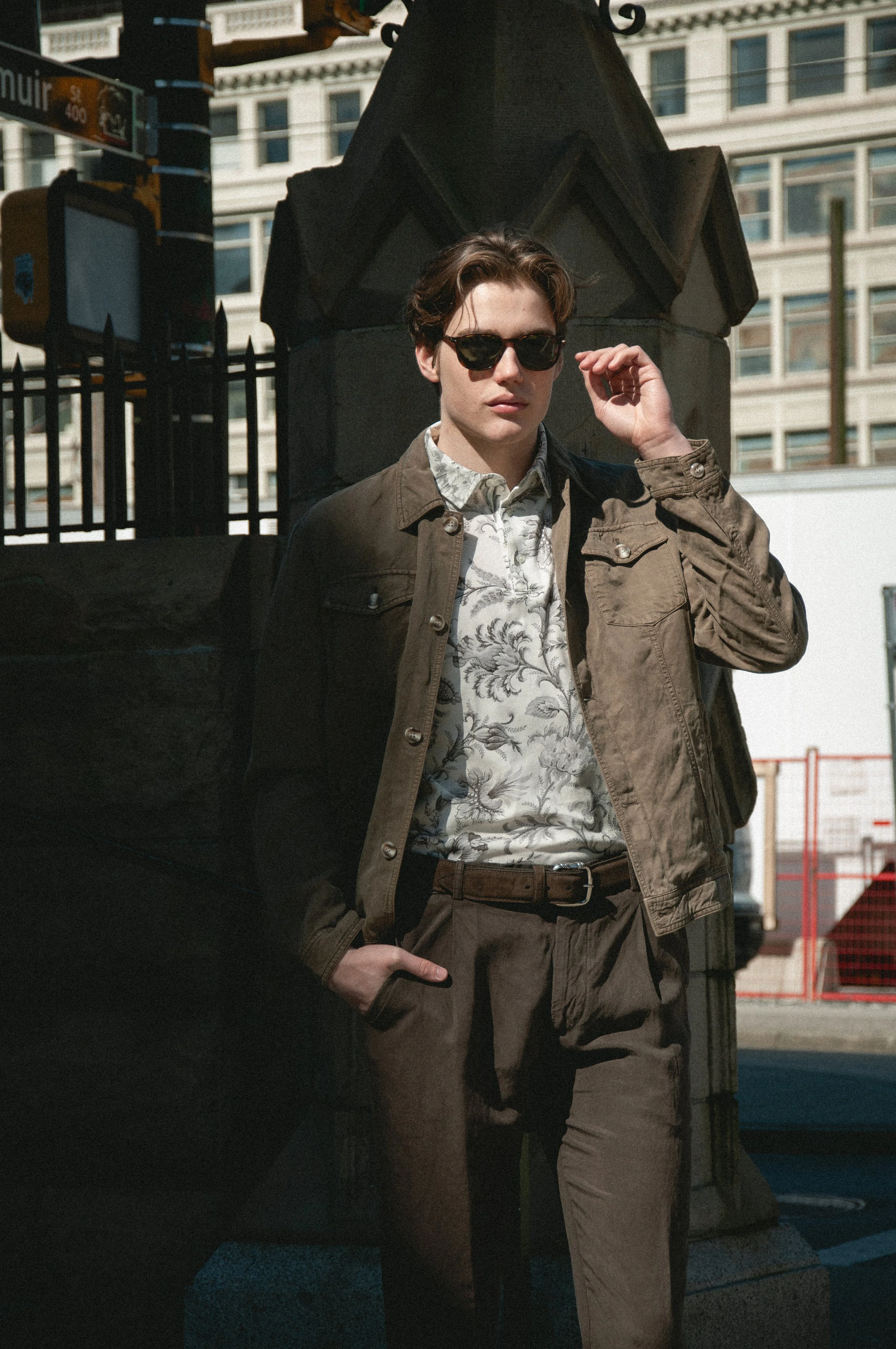 A young man with dark hair and sunglasses standing outdoors in front of a metal sculpture or monument. He is wearing a floral shirt, over which he has a brown jacket, and he has his hand near his face. Behind him are buildings with multiple windows, suggesting an urban environment.
