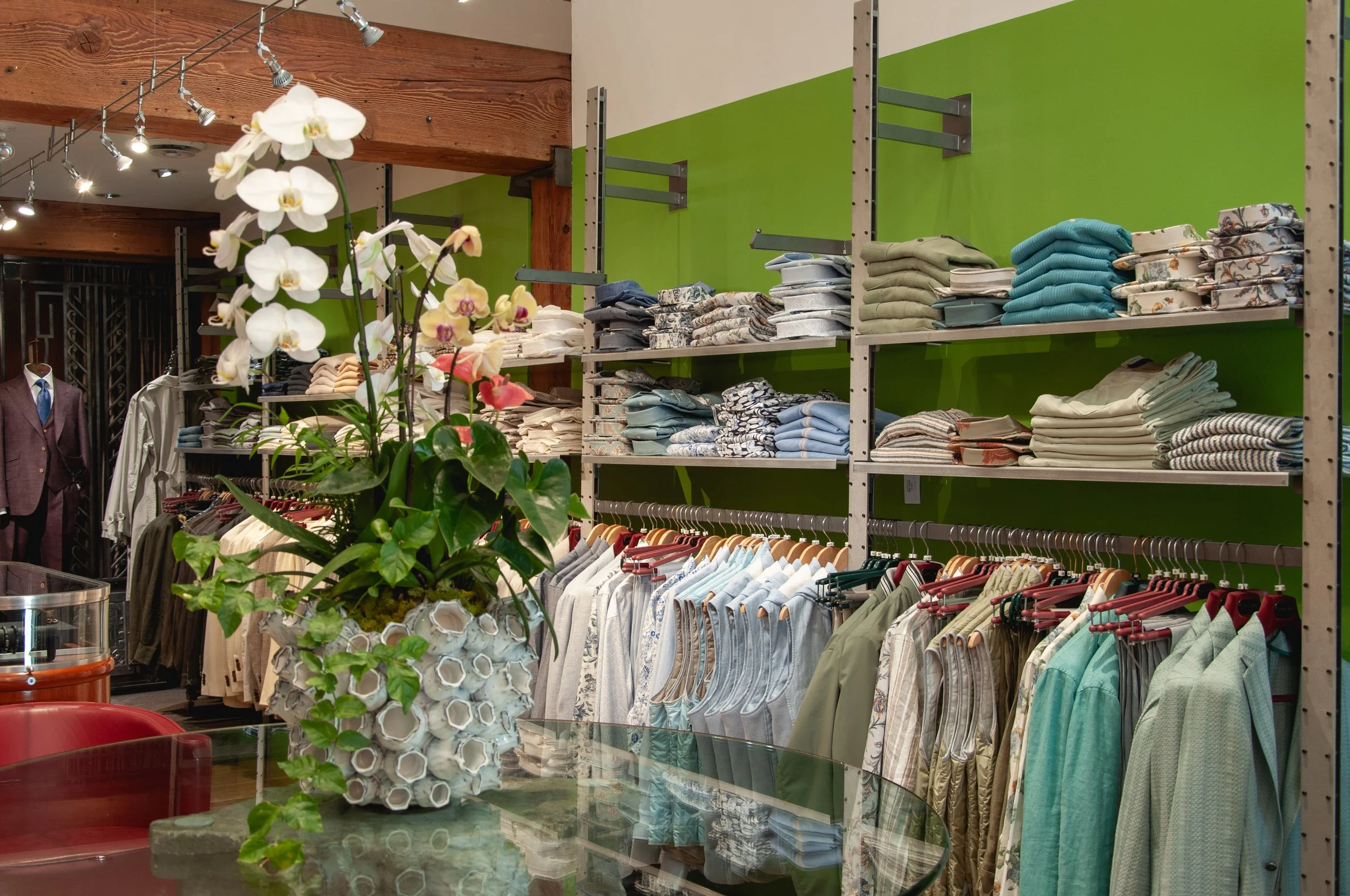 Clothing store display with shelves of neatly folded shirts and pants, a green painted wall, wooden beams on the ceiling, and a table with a floral arrangement of white orchids in the foreground.