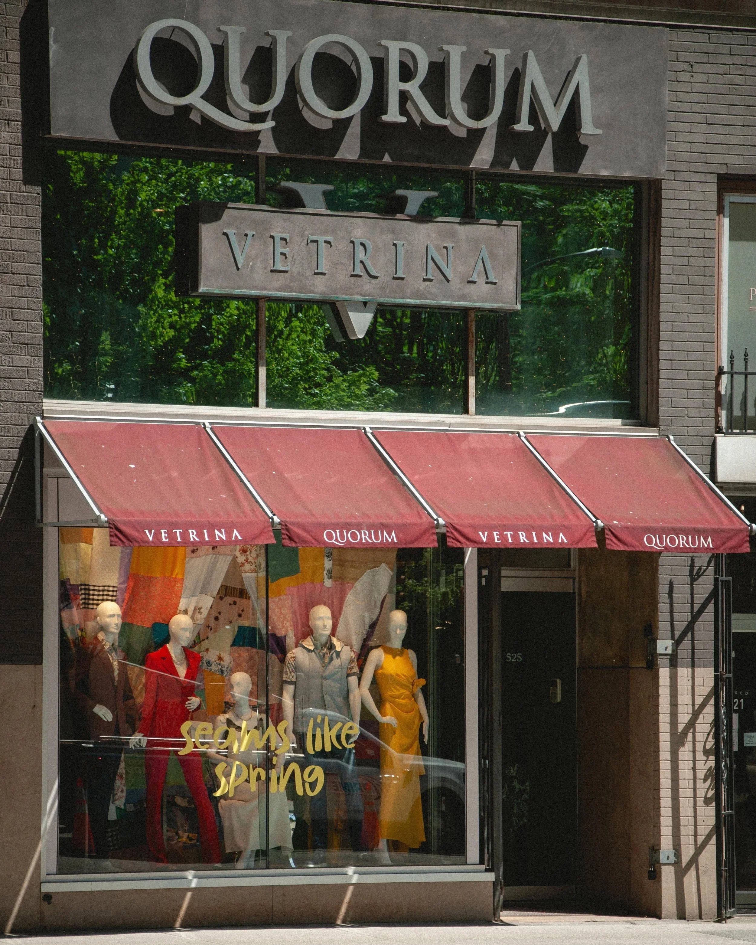 Store window display with mannequins dressed in colorful clothing and a sign that reads "Seams like Spring."" Above the window, there is a sign that says "QUORUM VETRINA" with the reflection of trees in the glass.