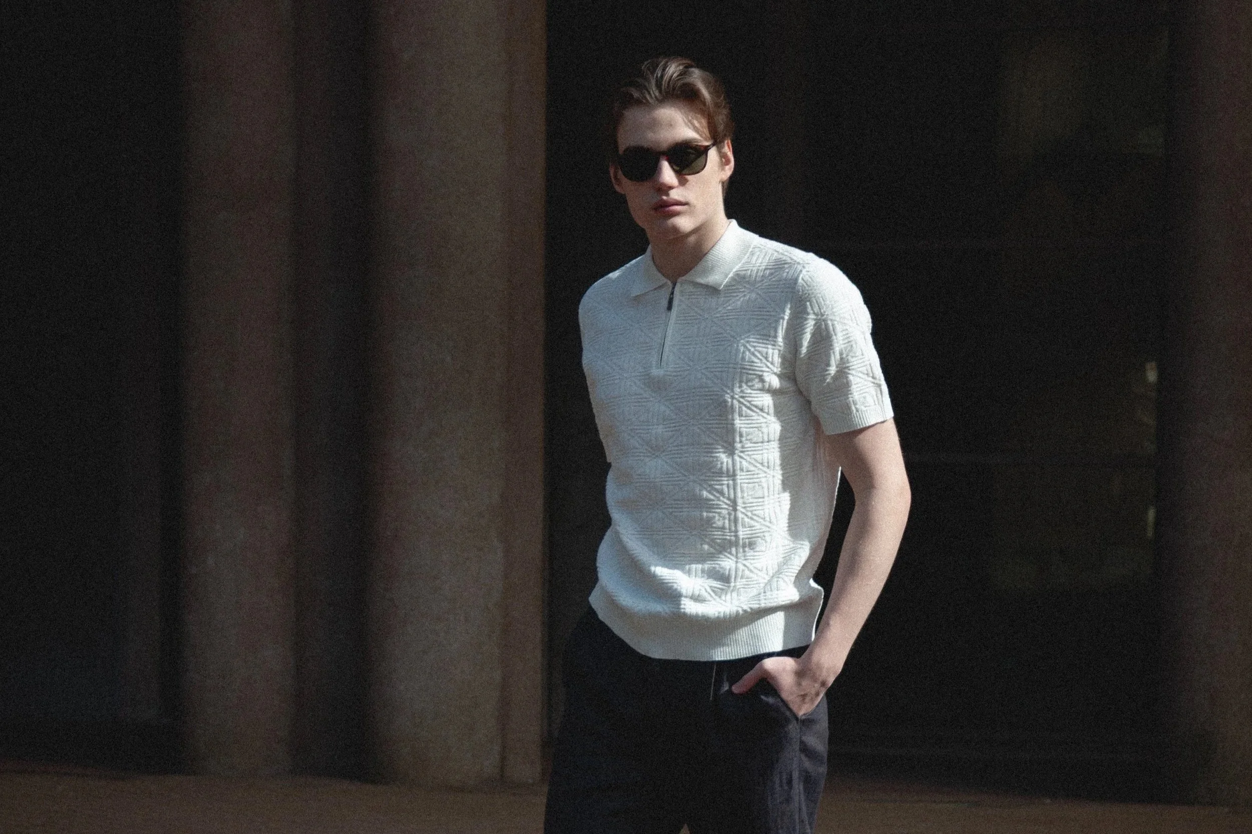 A young man with dark hair, wearing sunglasses, a white textured short-sleeve polo shirt, and dark pants, standing outdoors near two large columns.