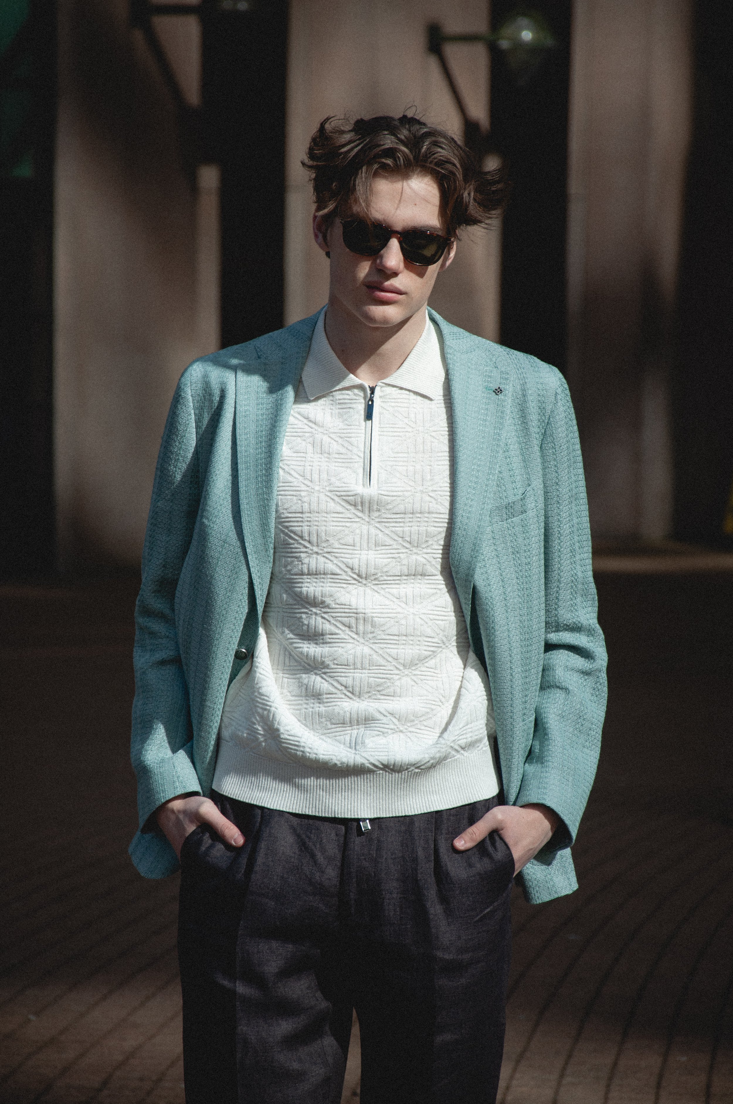 A young man with wavy hair wearing dark sunglasses, a light blue blazer, a white textured zip-up top, and dark plaid pants, standing outdoors with hands in pockets in front of a building.