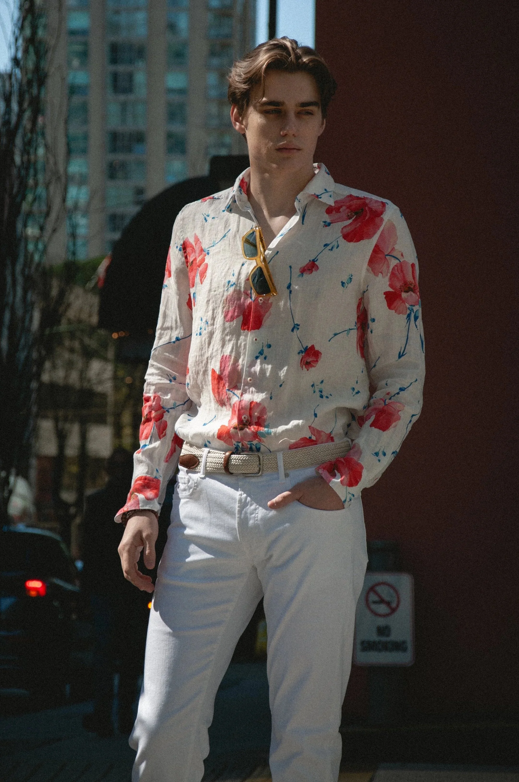 A young man with brown hair wearing a white shirt with a red floral pattern, white pants, and yellow sunglasses hanging on his shirt is standing outdoors on a sunny day.