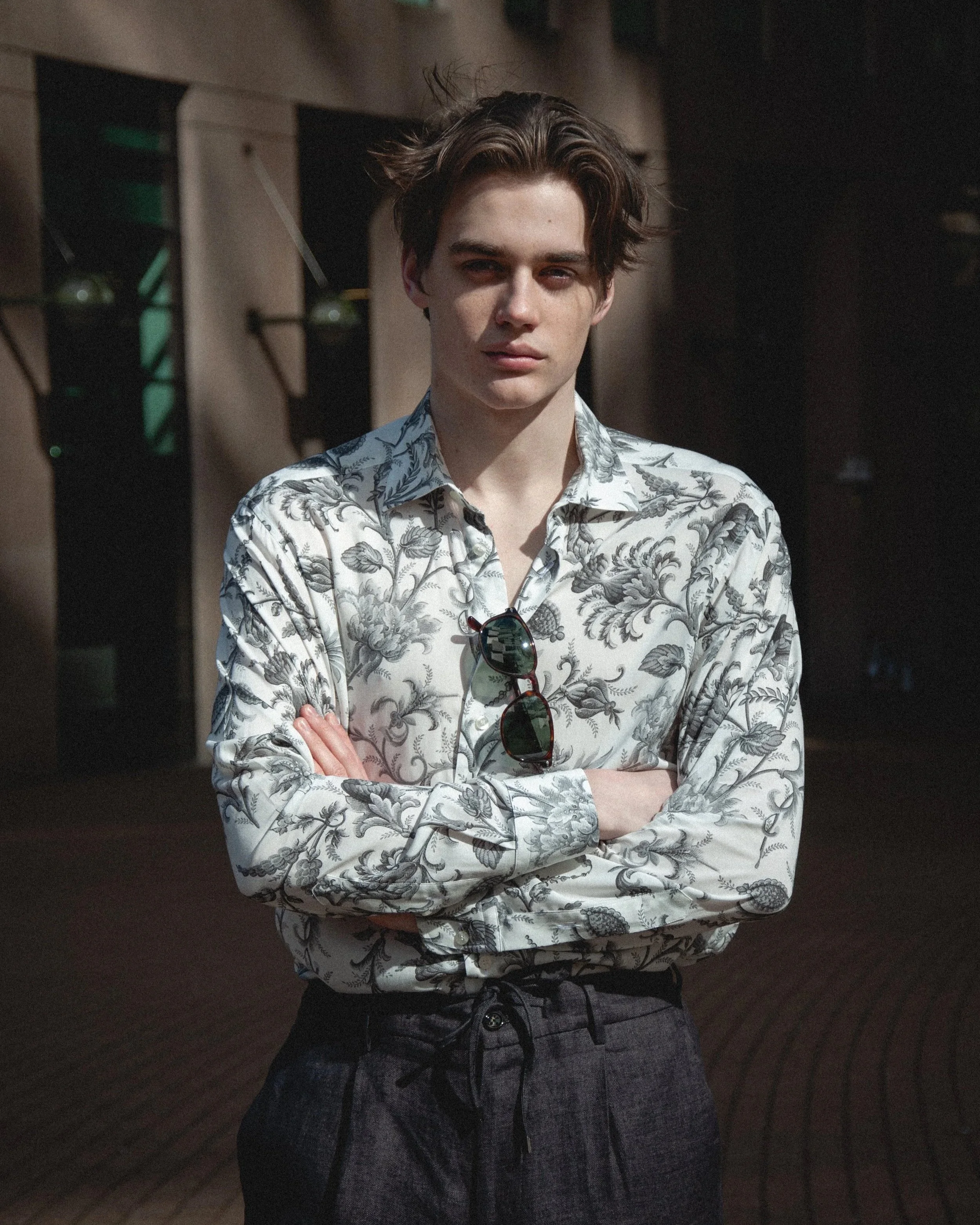 Young man with arms crossed, wearing a white shirt with gray floral pattern and gray shorts, standing in a dimly lit room.