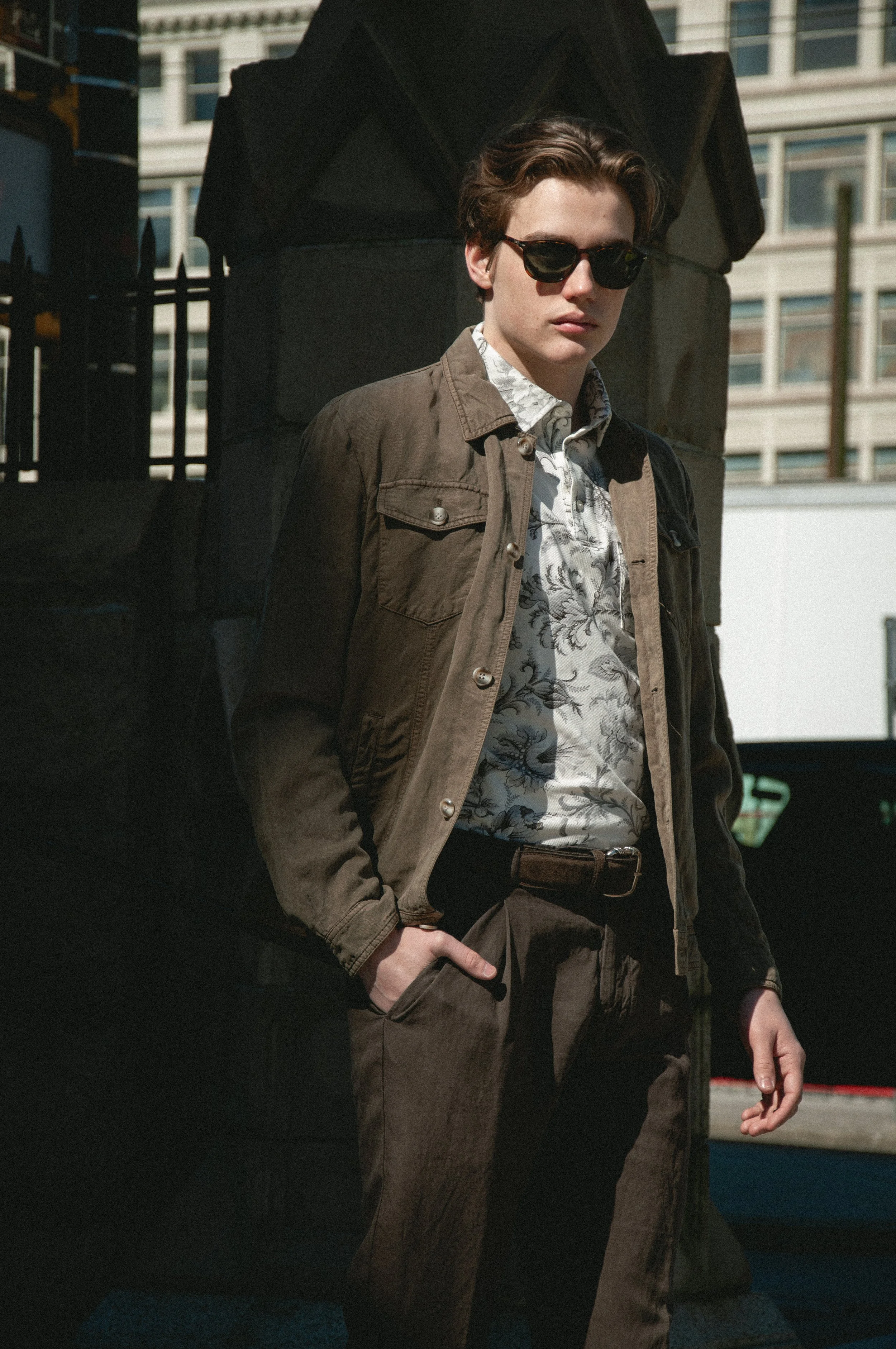 A young man with dark sunglasses, wearing a patterned shirt, brown jacket, and brown pants, standing outdoors in an urban setting near a black fence and large buildings in the background.