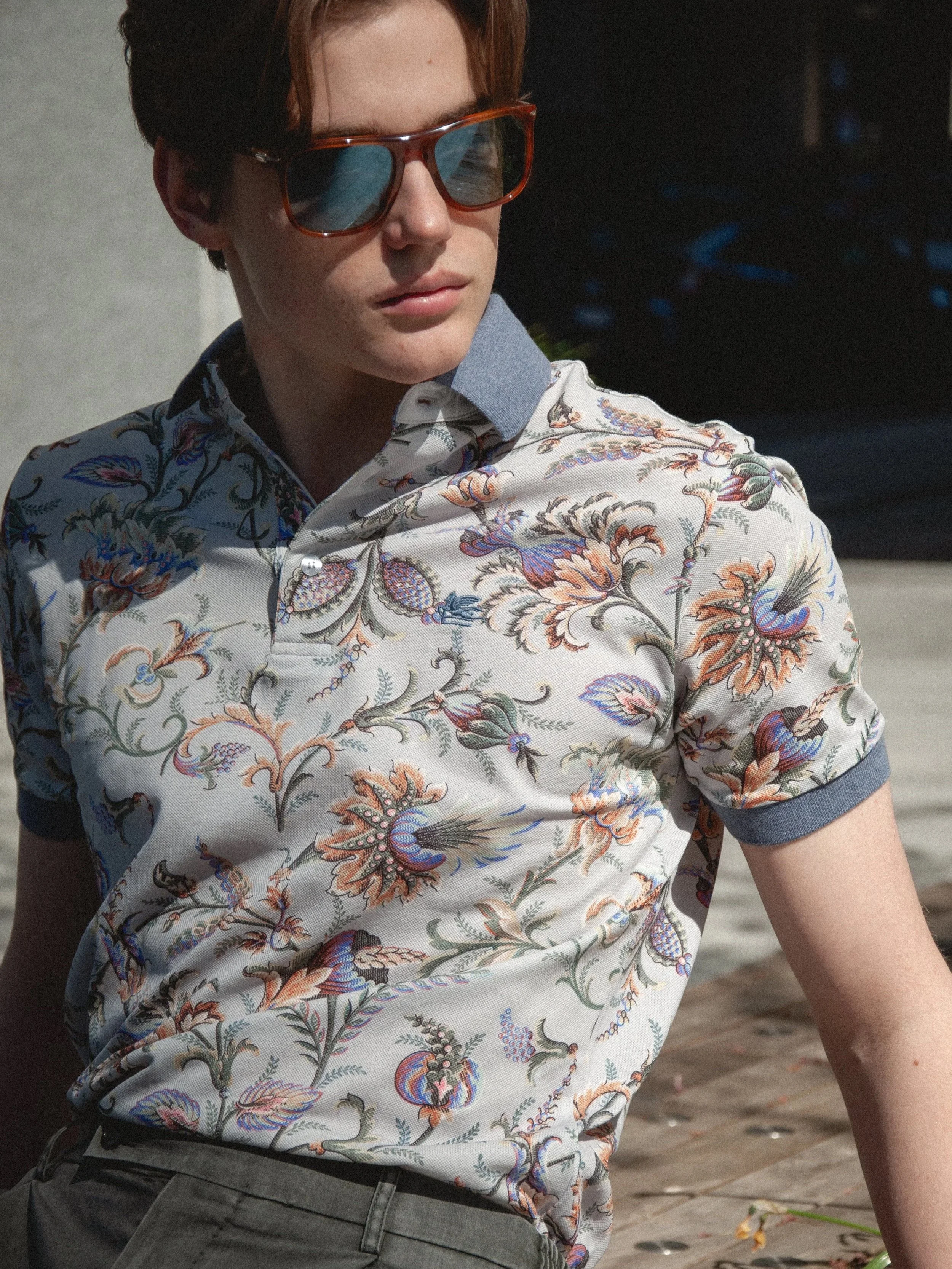 Young man with red hair, wearing sunglasses and a colorful, patterned short-sleeve shirt, standing outdoors on a wooden deck.