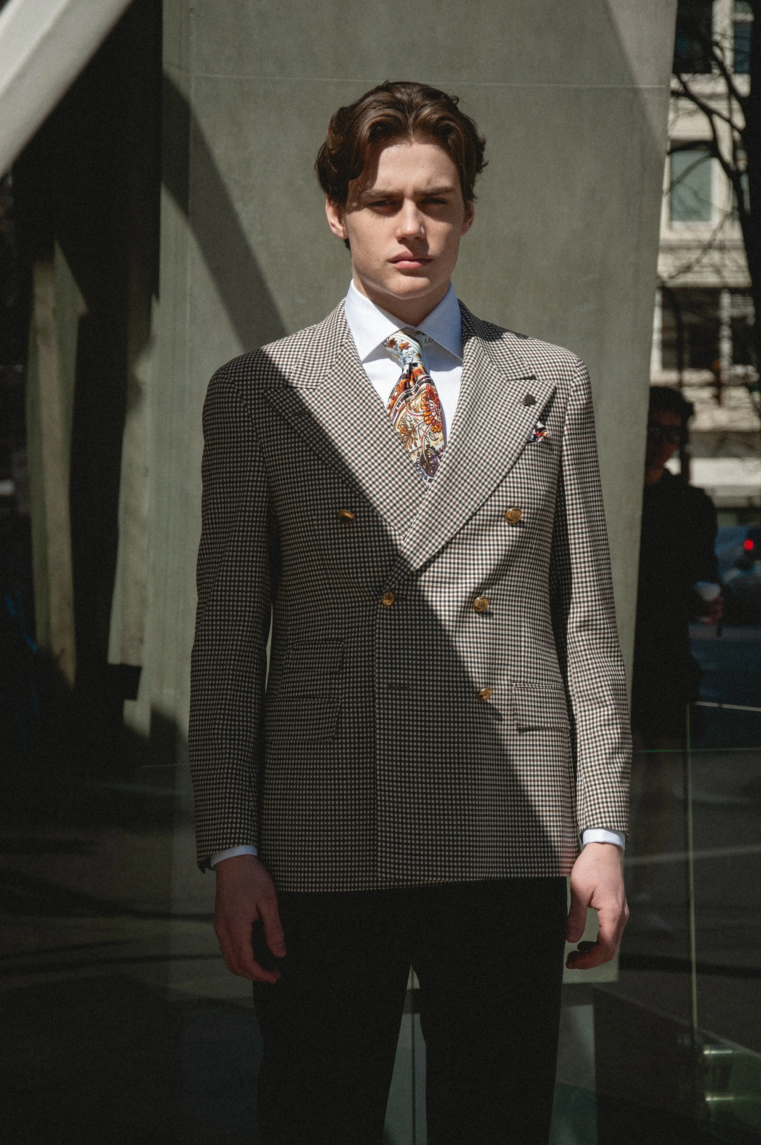 A young man with brown hair standing outdoors in a checkered brown blazer, white shirt, and colorful tie, with a serious expression.
