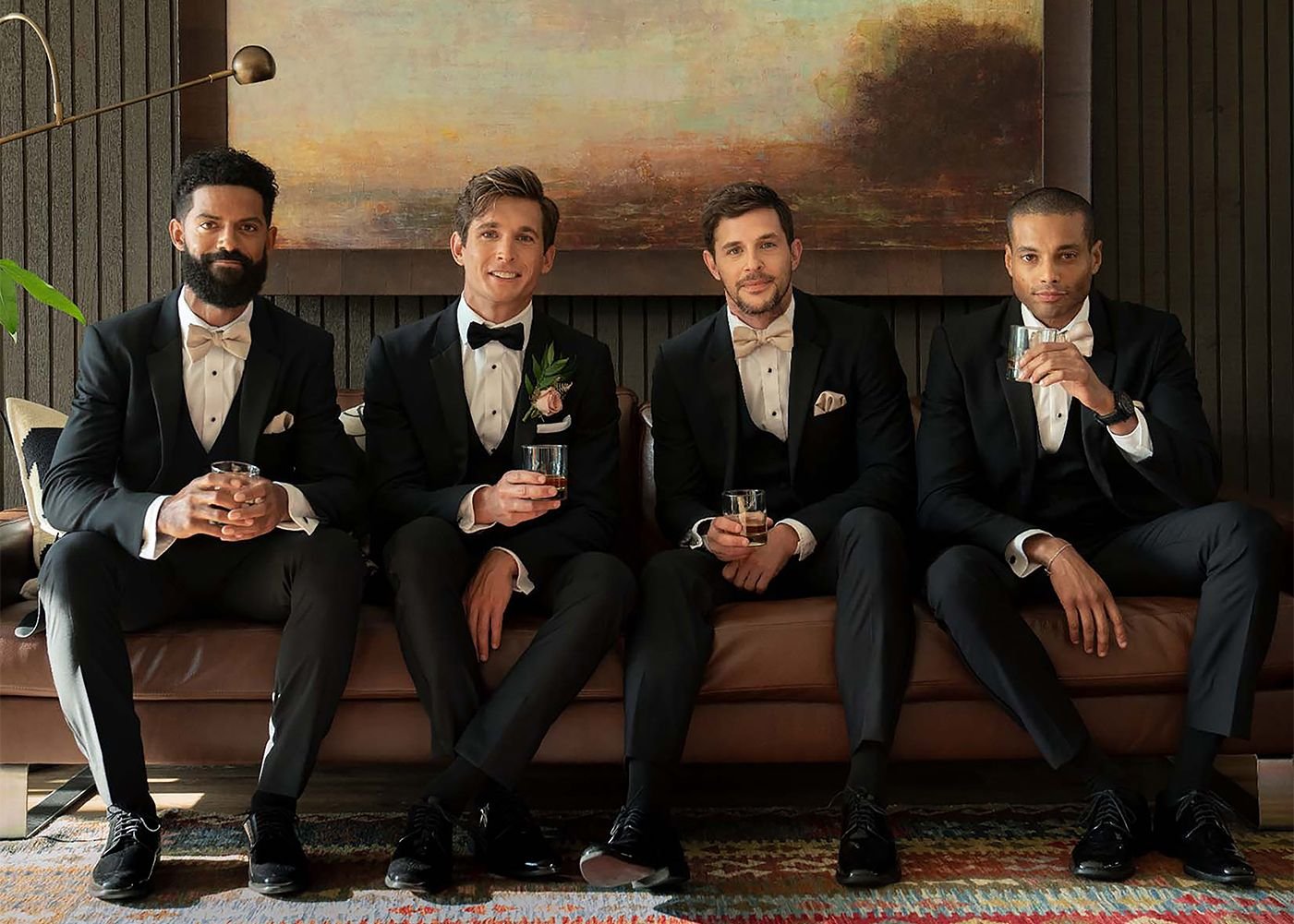 Four men dressed in black tuxedos with white shirts and bow ties, sitting on a sofa and holding drinks at a formal event.