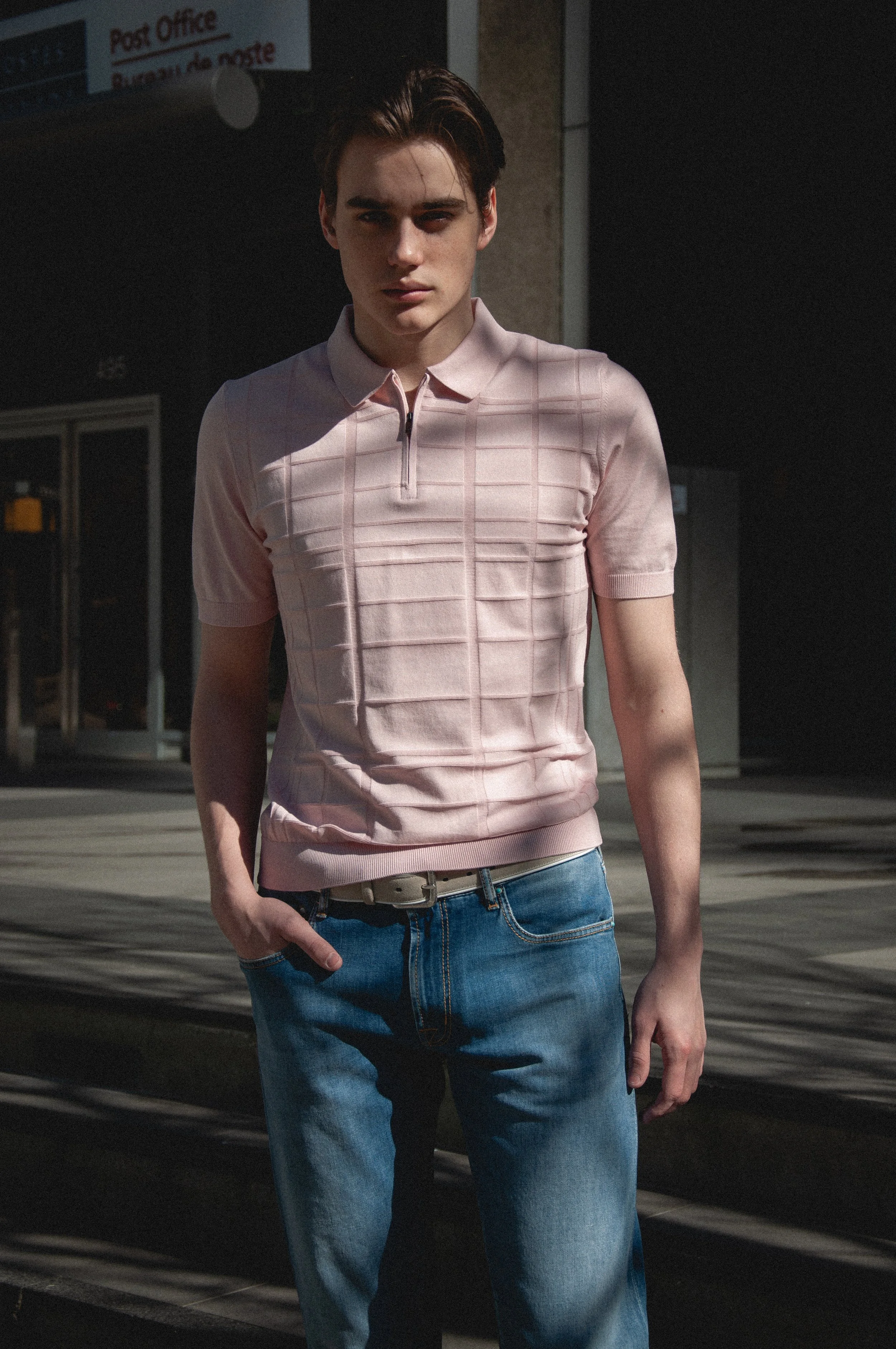 A young man with dark hair, wearing a pink plaid short-sleeve shirt and blue jeans, standing outdoors in front of a moving vehicle and a building, with a shadow cast across his face.