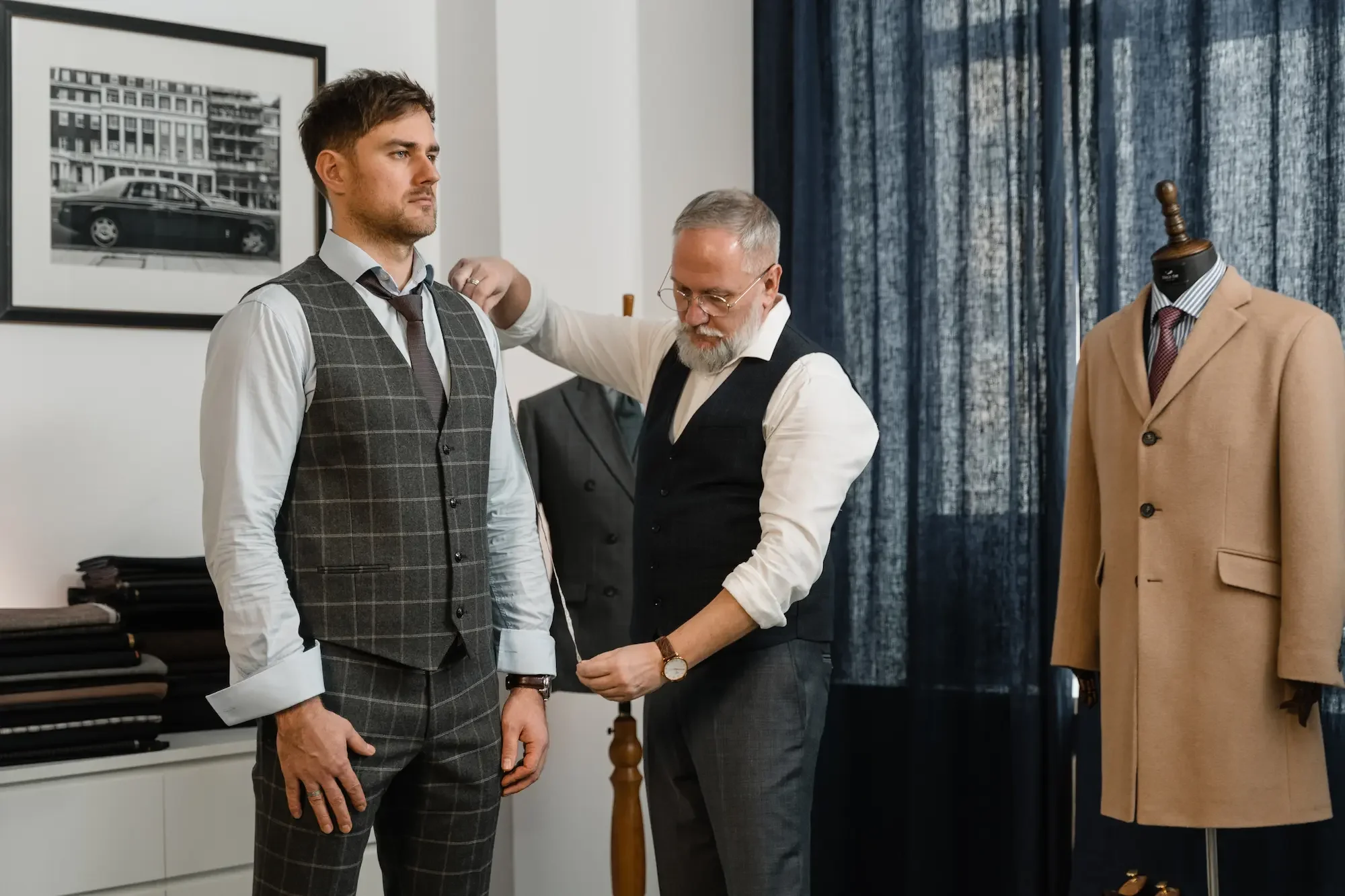 A tailor fitting a suit on a male customer in a clothing store, with other suits and a photograph of a car and building on the wall in the background.