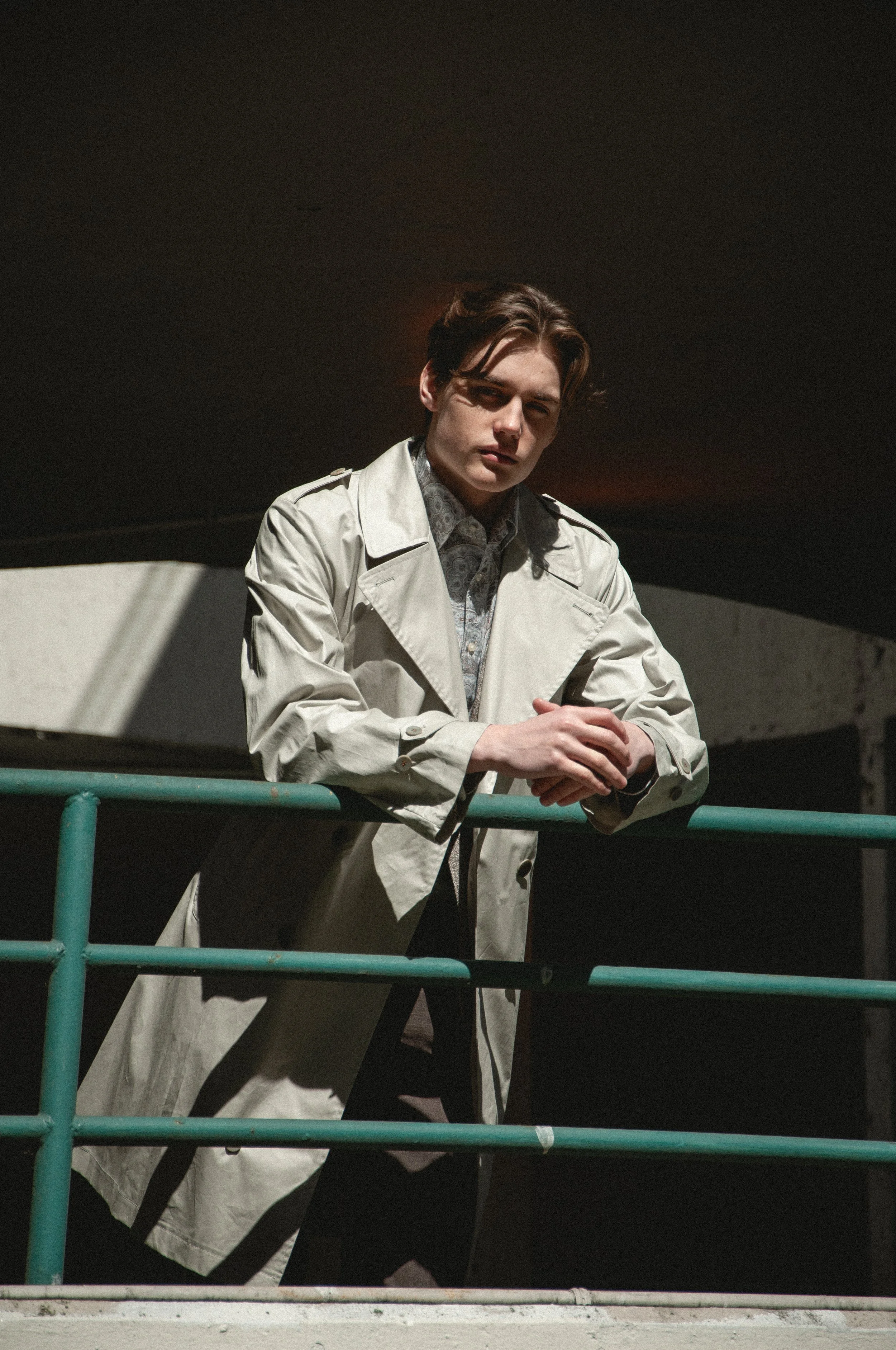 Young man with brown hair wearing a beige trench coat and patterned shirt, standing behind a green railing at night.