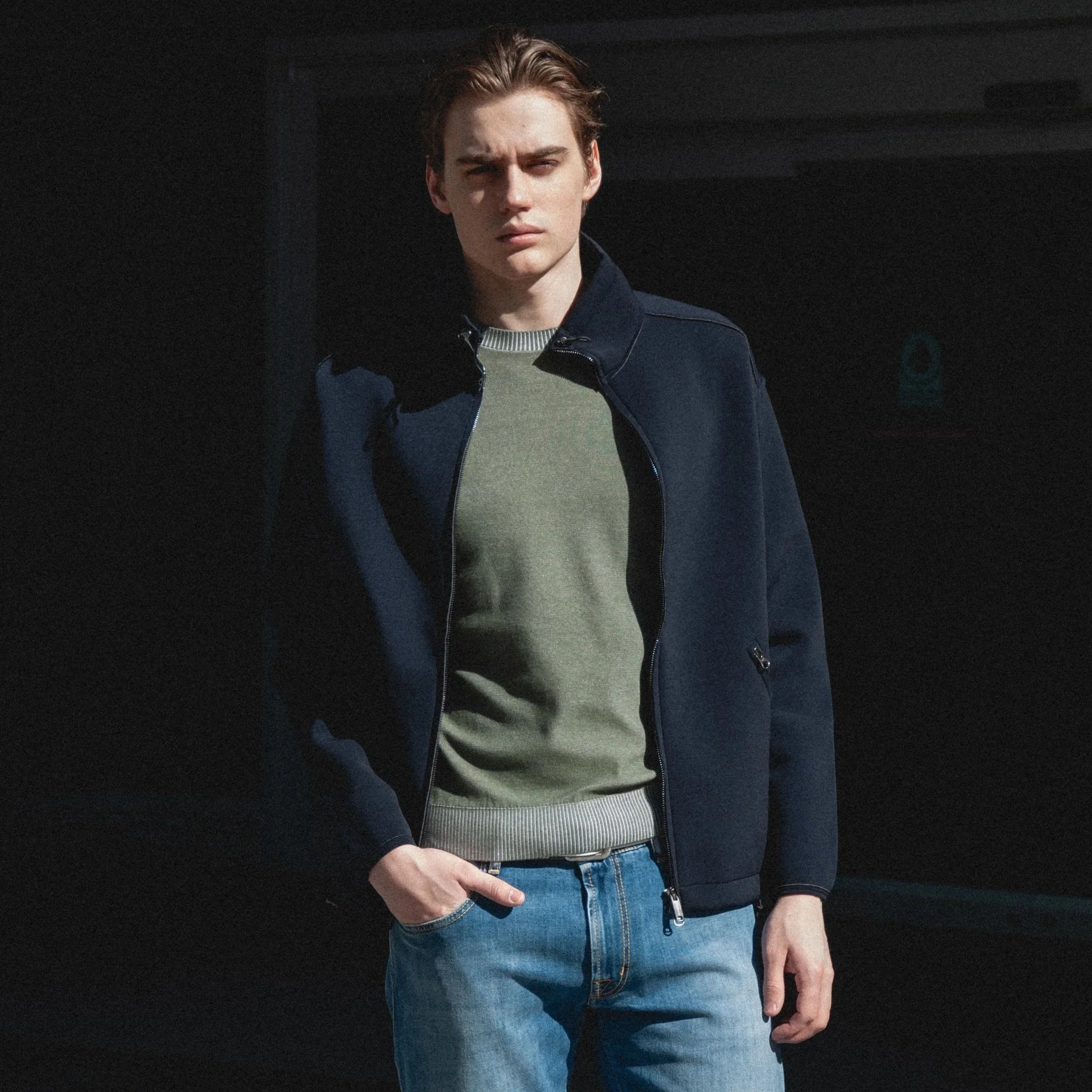 A young man with reddish-brown hair, wearing a dark blue jacket over a light-colored sweater and blue jeans, making a peace sign with his right hand against a dark background.